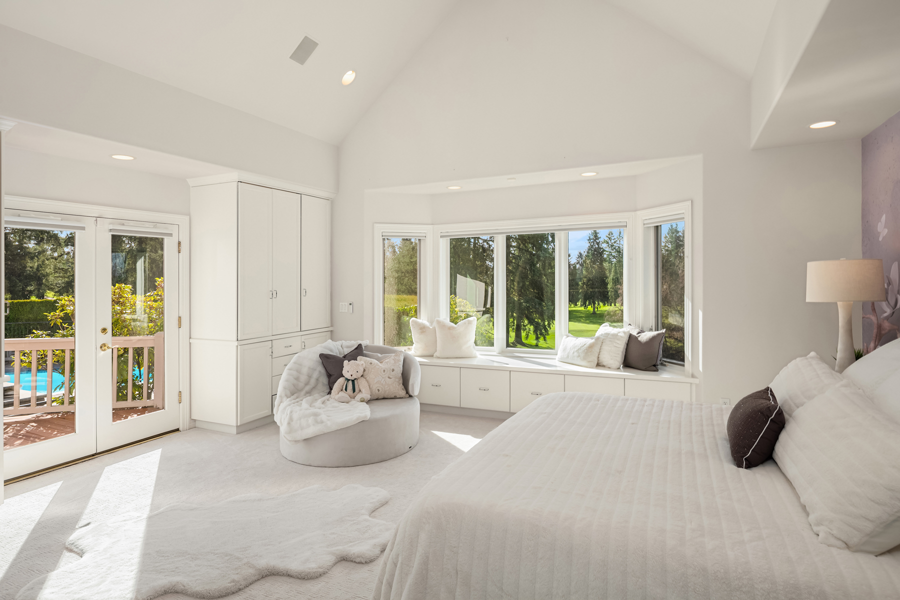 This is a bright and airy primary bedroom featuring a large bay window with a built-in bench seat, offering views of the lush greenery outside. The room is decorated in a neutral color palette with white walls, carpet, and furniture, creating a serene and inviting atmosphere. A cozy armchair and a plush rug add to the comfort of the space, while French doors lead to an outdoor balcony.