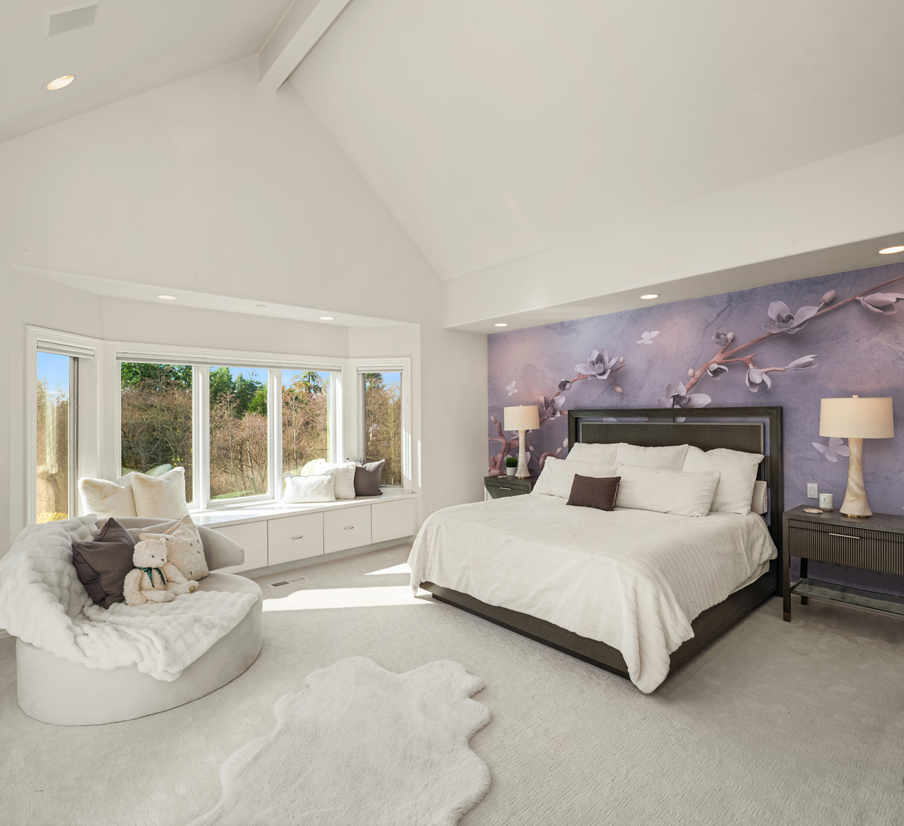 This is a bright and spacious primary bedroom featuring a king-size bed with white linens and decorative pillows, set against a floral-patterned accent wall. A bay window with a built-in bench provides natural light and a cozy seating area. The room is carpeted in a neutral tone, creating a serene and inviting atmosphere.