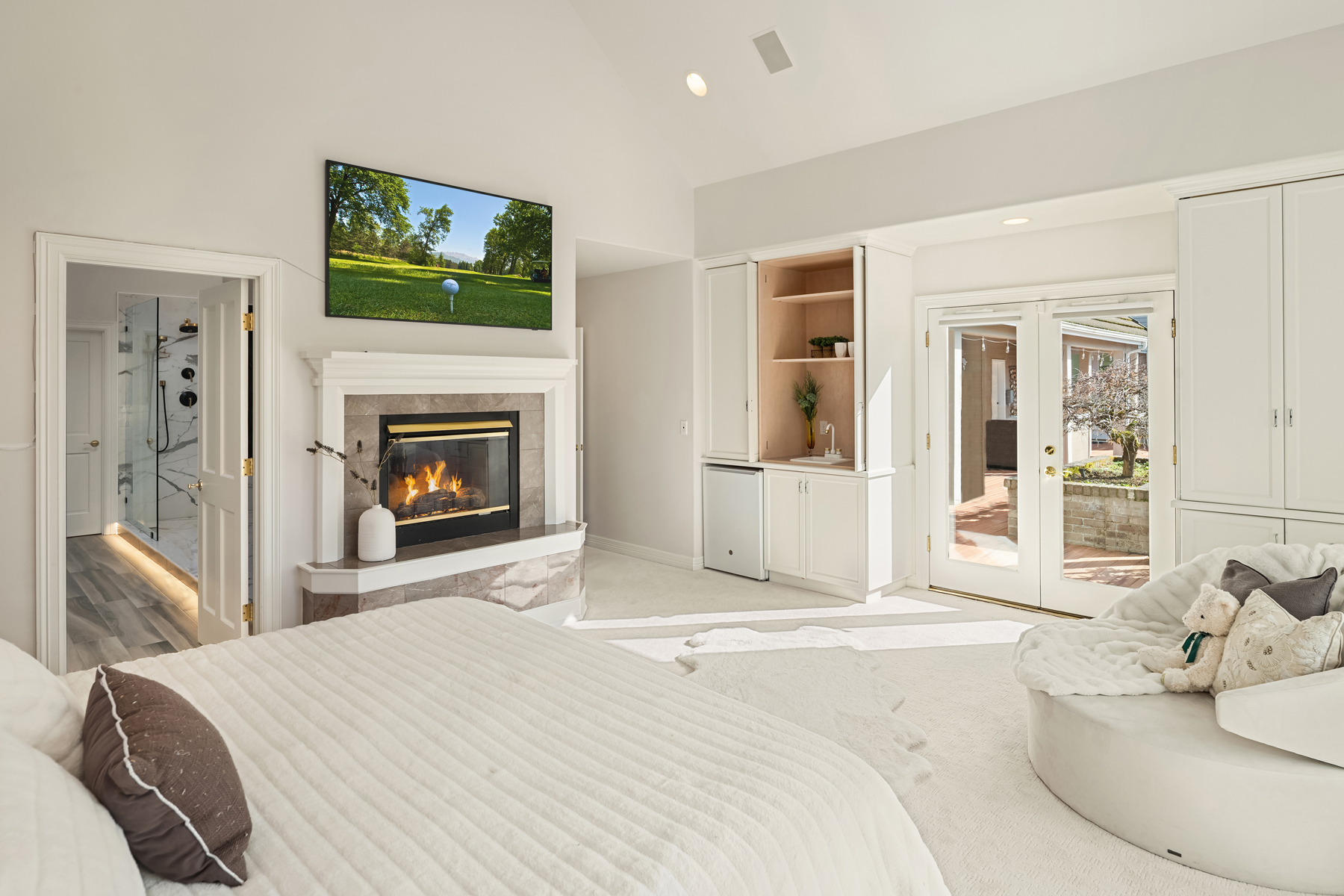 This is a bright and spacious primary bedroom featuring a fireplace with a TV mounted above it. The room has a luxurious feel with a large bed, a cozy round chair, and access to an outdoor patio through French doors. Built-in cabinetry provides ample storage, and the overall color scheme is light and airy.