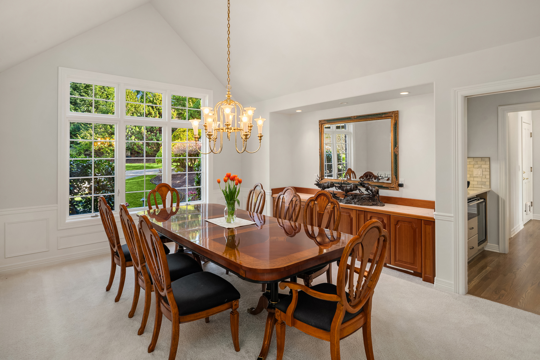 This is an interior shot of a formal dining room. The room features a large wooden dining table with black cushioned chairs, a gold chandelier, and a large window providing natural light. A wooden sideboard with a decorative mirror and sculpture adds to the room's elegance.