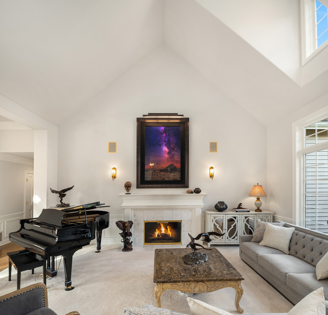 This is a spacious living room featuring a grand piano, a fireplace with a framed artwork above, and a comfortable-looking gray sofa. The room has high ceilings and is well-lit, creating an elegant and inviting atmosphere. The decor includes sculptures and a detailed coffee table, adding to the room's sophisticated style.