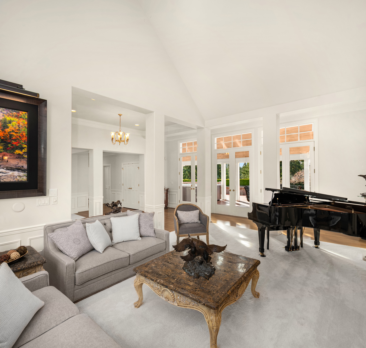 This is an interior shot of a luxurious living room featuring a grand piano, a comfortable-looking sofa with decorative pillows, and an ornate coffee table with a sculpture. The room is brightly lit with natural light streaming through large windows, and the neutral color palette creates a sophisticated and inviting atmosphere. The perspective is a wide shot, capturing the spaciousness and elegance of the room.