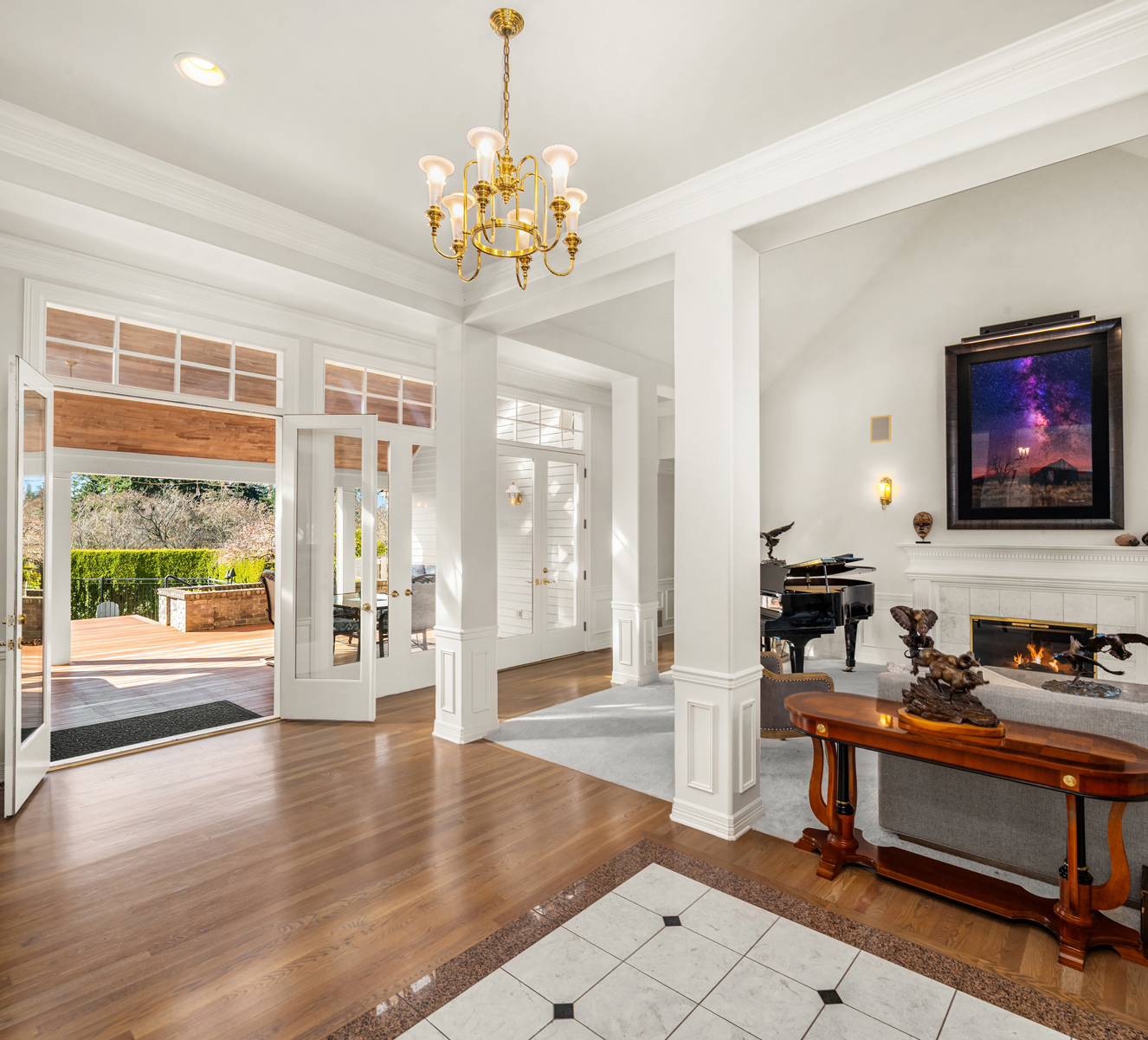 This grand living room features an open floor plan with hardwood flooring transitioning to tile at the entrance. The space is illuminated by a gold chandelier and natural light streaming through large windows and French doors leading to an outdoor deck. A fireplace with a decorative mantle, a grand piano, and elegant furnishings contribute to the room's luxurious and inviting atmosphere.
