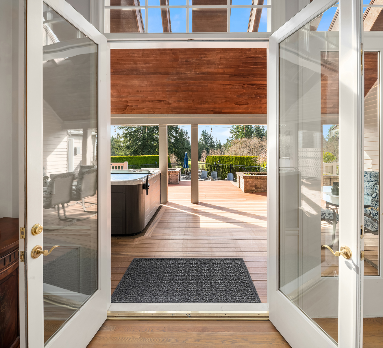 The image showcases an inviting outdoor living space, viewed through open French doors. A spacious wooden deck features a hot tub, lounge chairs, and a well-maintained hedge providing privacy. The covered portion of the deck has a wood ceiling, creating a cozy and stylish transition between indoor and outdoor living.