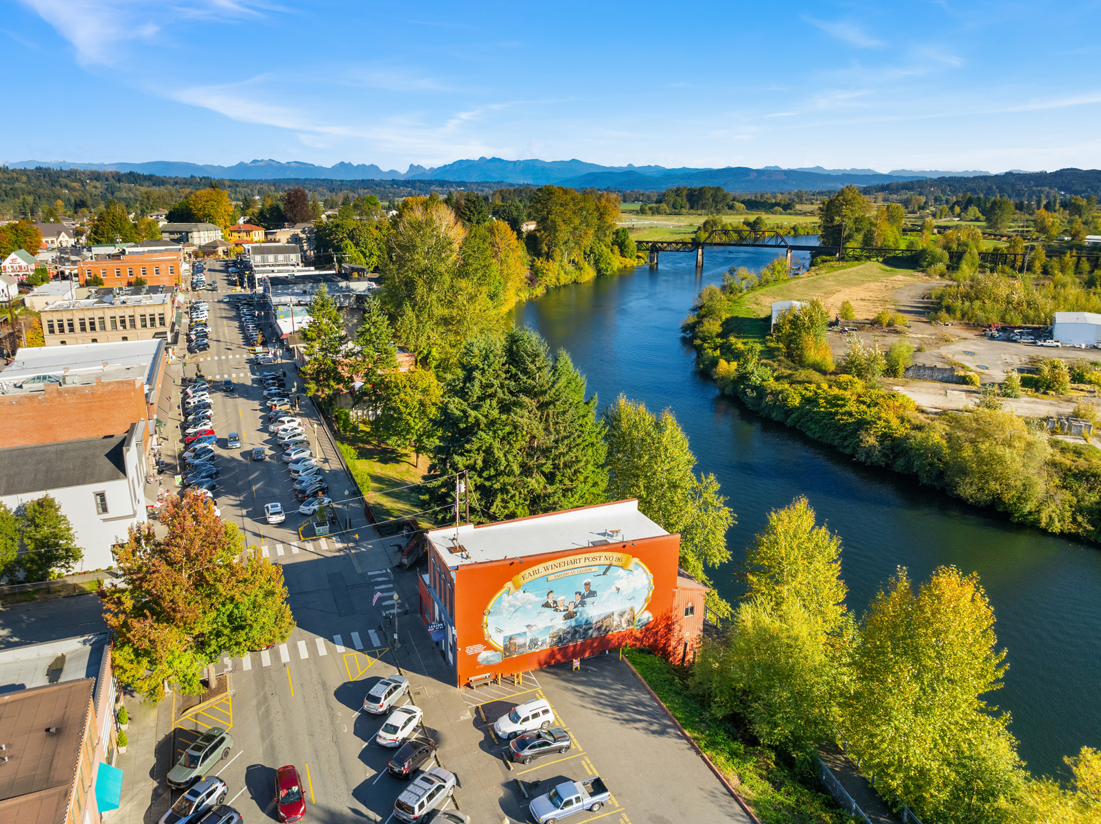 This aerial view showcases a charming town nestled alongside a river, with a bridge connecting both sides. The buildings are a mix of brick and other materials, with cars lining the streets, indicating a vibrant community. Lush greenery and trees surround the town, with mountains visible in the distance, creating a picturesque and inviting atmosphere.