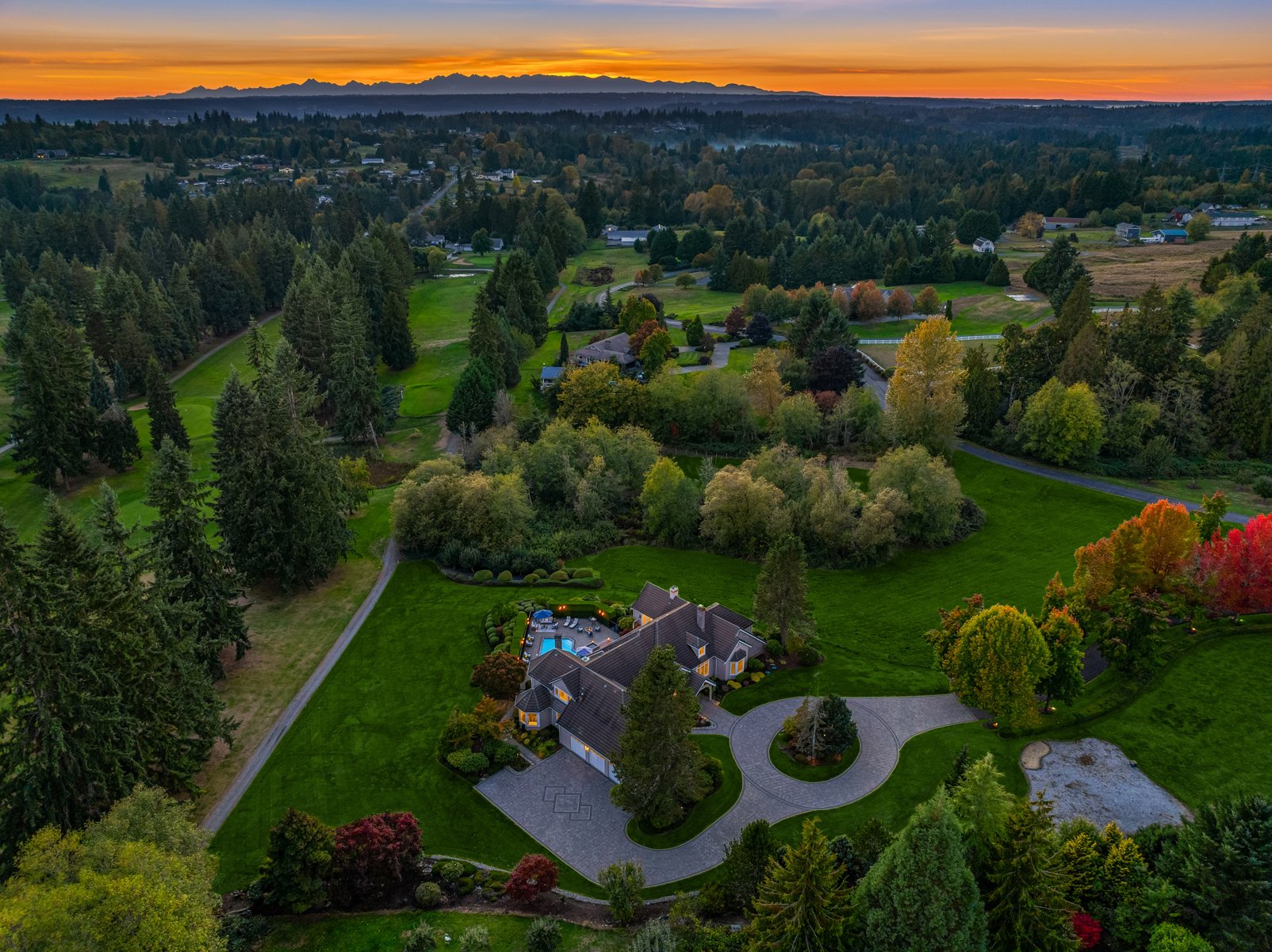 This aerial view showcases a luxurious estate nestled within lush greenery. The property features a large house with a circular driveway, a swimming pool, and meticulously landscaped gardens. The surrounding area includes a golf course and mature trees, creating a sense of privacy and exclusivity, with a beautiful sunset in the background.