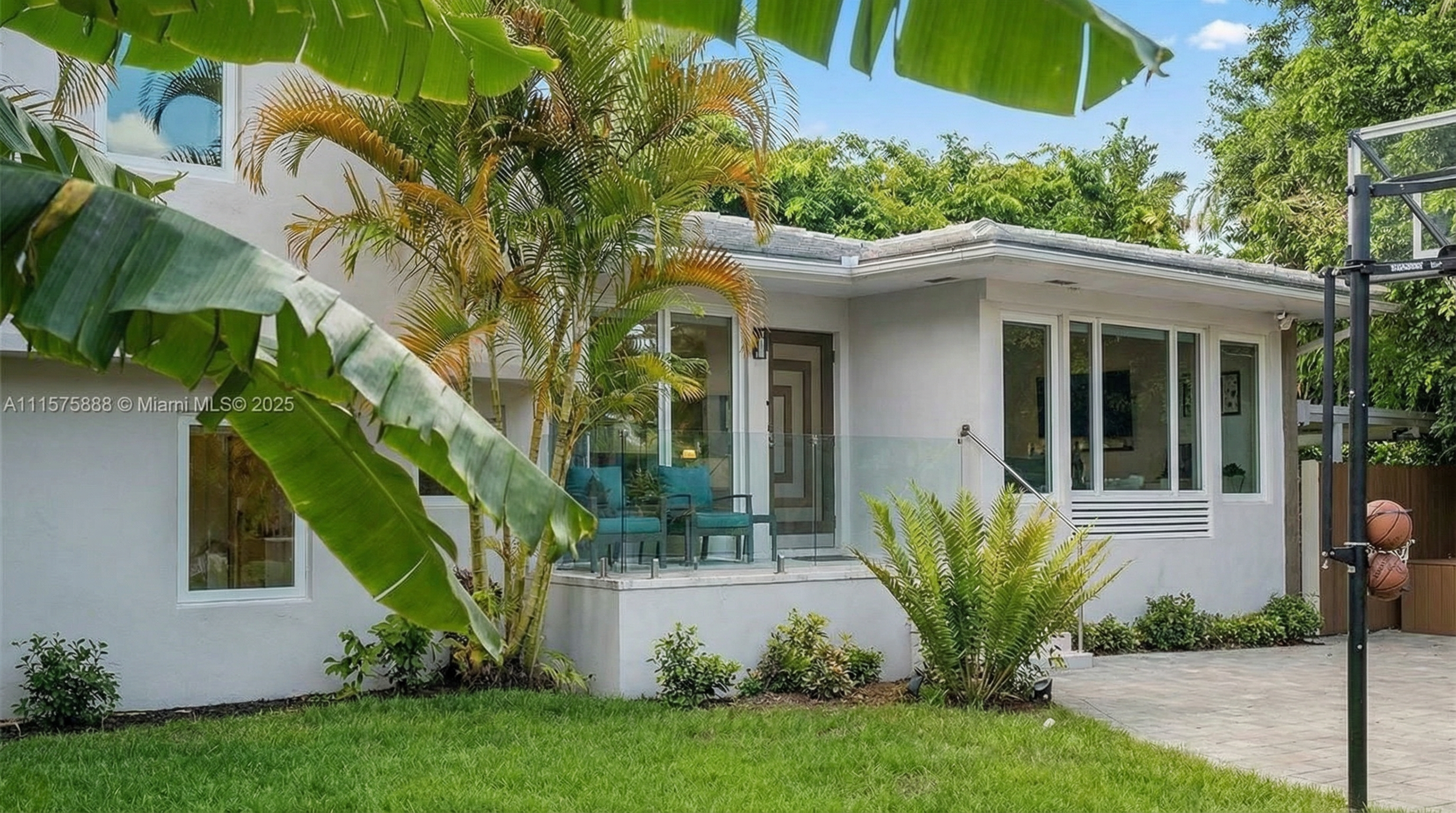 The image showcases the front exterior of a modern home with lush tropical landscaping. The house features a light gray facade, large windows providing ample natural light, and a small patio area with seating. A basketball hoop is visible on the side, suggesting recreational space.