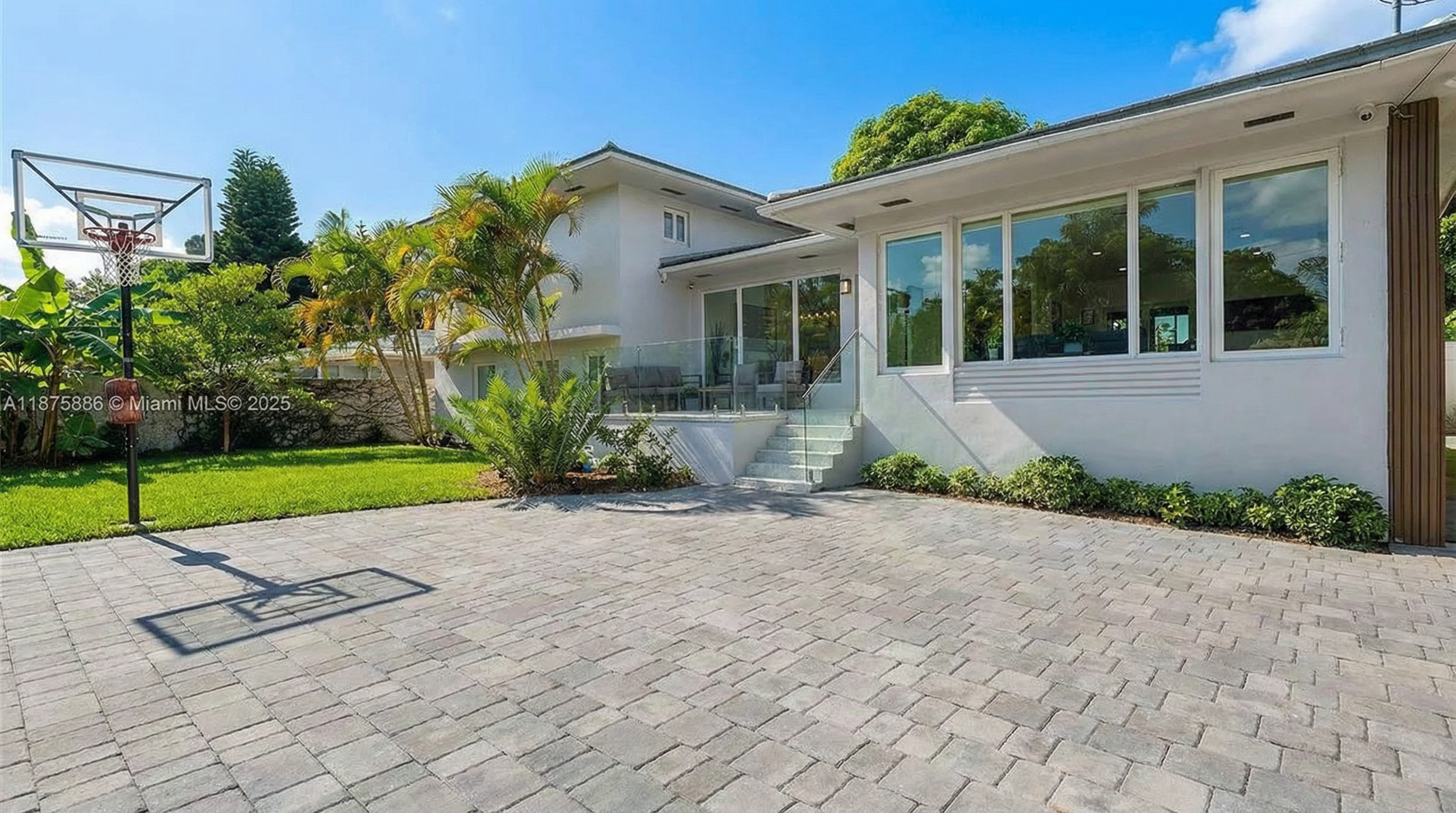 This image showcases the rear exterior of a modern residence. The property features a paved patio, lush landscaping, and glass-enclosed extensions that highlight the contemporary architectural design. A basketball hoop is also visible, adding an element of recreation to the outdoor space.