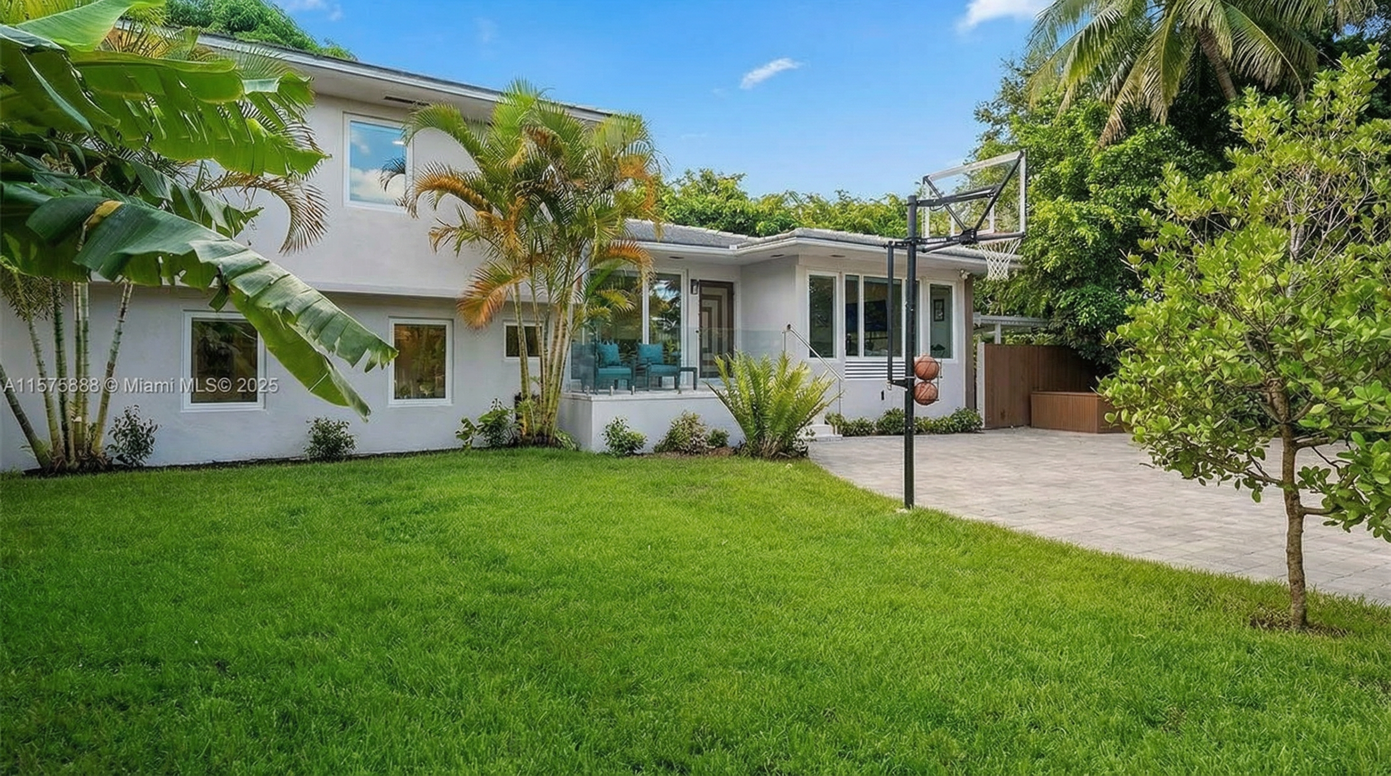 The image showcases the front exterior of a modern two-story home with light-colored stucco. Lush green lawn and manicured landscaping add to the curb appeal. A basketball hoop is present in the driveway, suggesting a family-friendly environment.