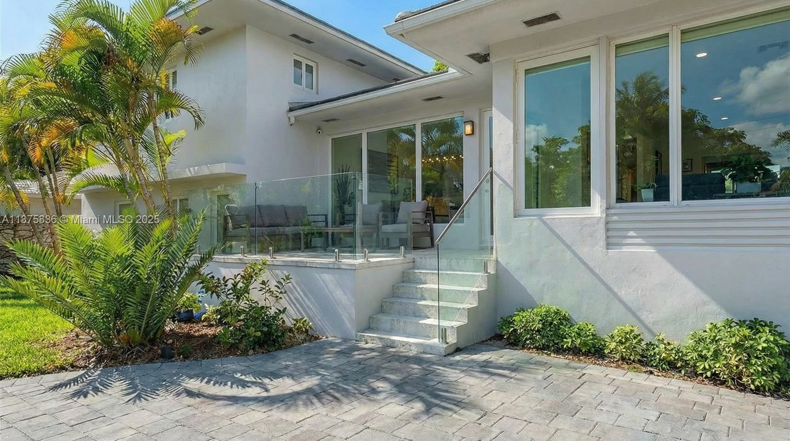 The image showcases an exterior patio or deck area of a modern home. It features glass railings, light-colored stone steps, and a paved patio surface. The outdoor space is bordered by lush greenery, offering a sense of privacy and a connection to nature, enhancing the property's appeal for outdoor living.