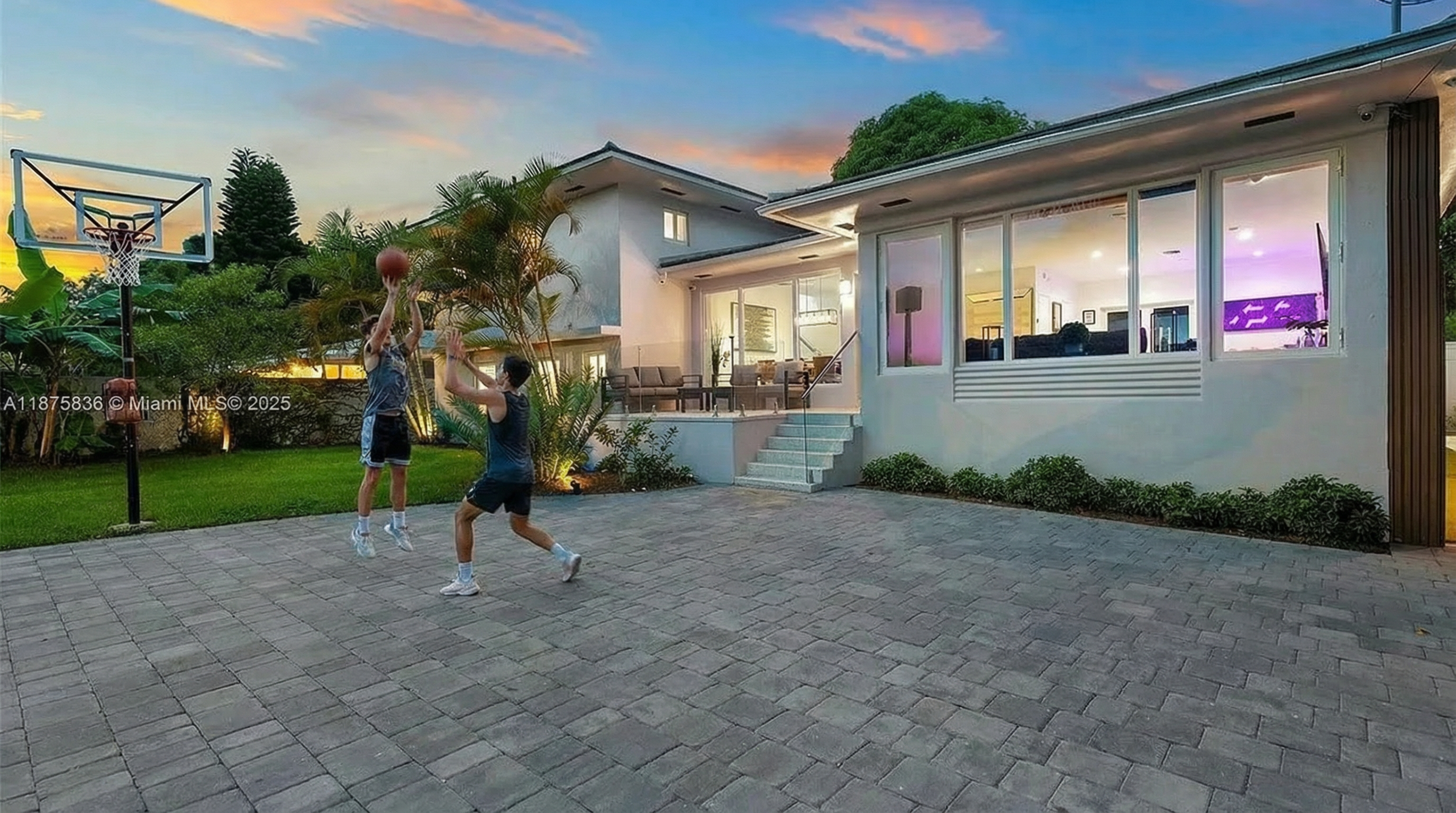 This image showcases the rear of a modern residence with a basketball court in the backyard. The property features a contemporary architectural style with large windows that offer views into the interior. The outdoor area is designed for recreation and entertaining.