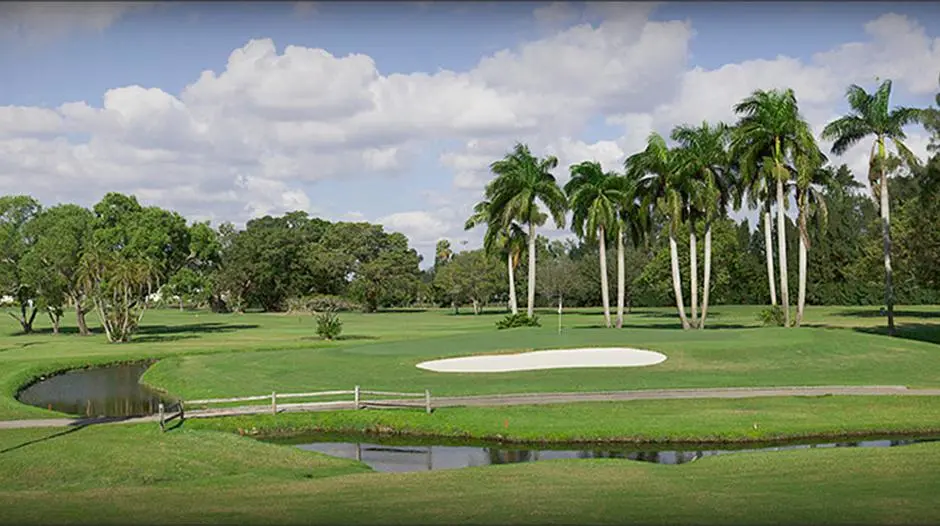 The image portrays a well-manicured golf course, likely part of a community's amenities. A sand trap is visible near the green, and a small water feature with a wooden bridge adds to the visual appeal. Palm trees dot the landscape under a partly cloudy sky, contributing to a lush, inviting atmosphere.