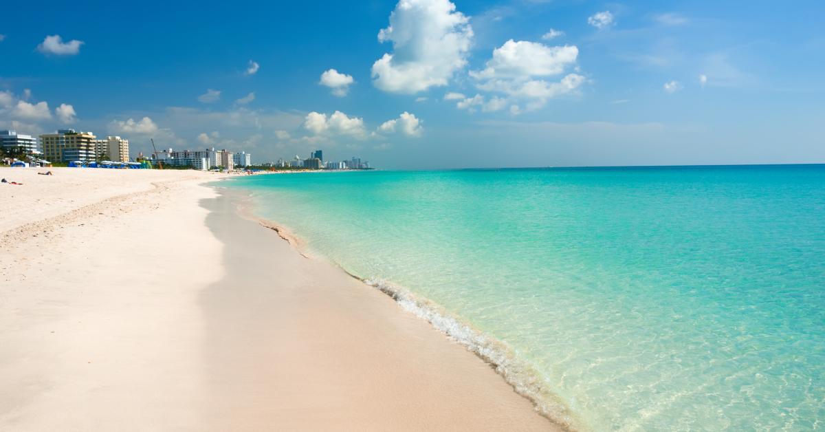 This is a scenic shot of a beachfront property showcasing a beautifully maintained white sand beach and clear turquoise water. In the background, you can see a few residential and hotel buildings suggesting the property is located near coastal amenities. The overall atmosphere is inviting and conveys luxury and relaxation.