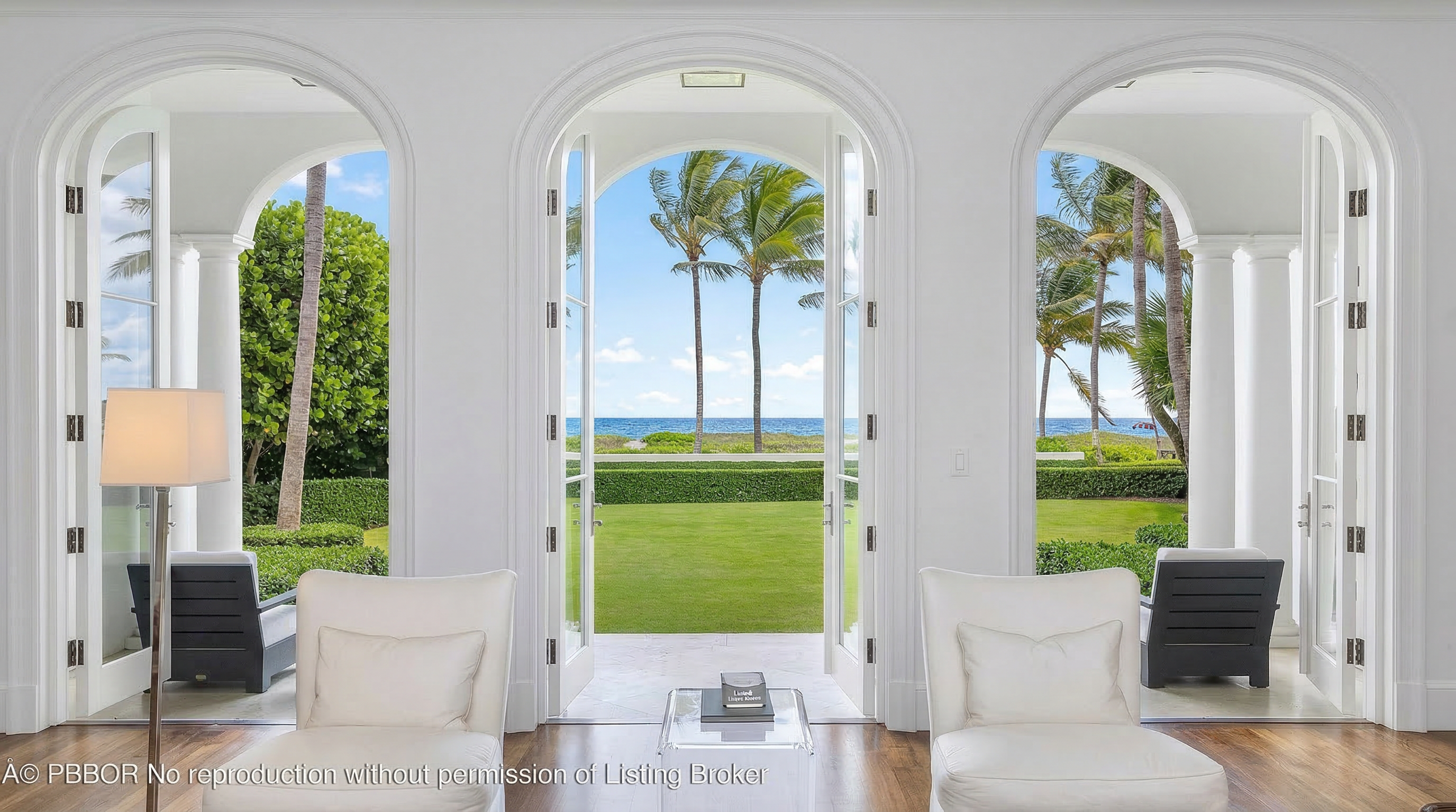 This elegant interior space features three symmetrical floor-to-ceiling arched doorways that frame a stunning view of a private lawn, palm trees, and the ocean beyond. The room is decorated with minimalist white armchairs and a modern lamp, emphasizing a sophisticated and airy aesthetic typical of a luxury coastal home. The seamless transition between the indoor living area and the outdoor landscape creates an expansive, resort-like atmosphere.