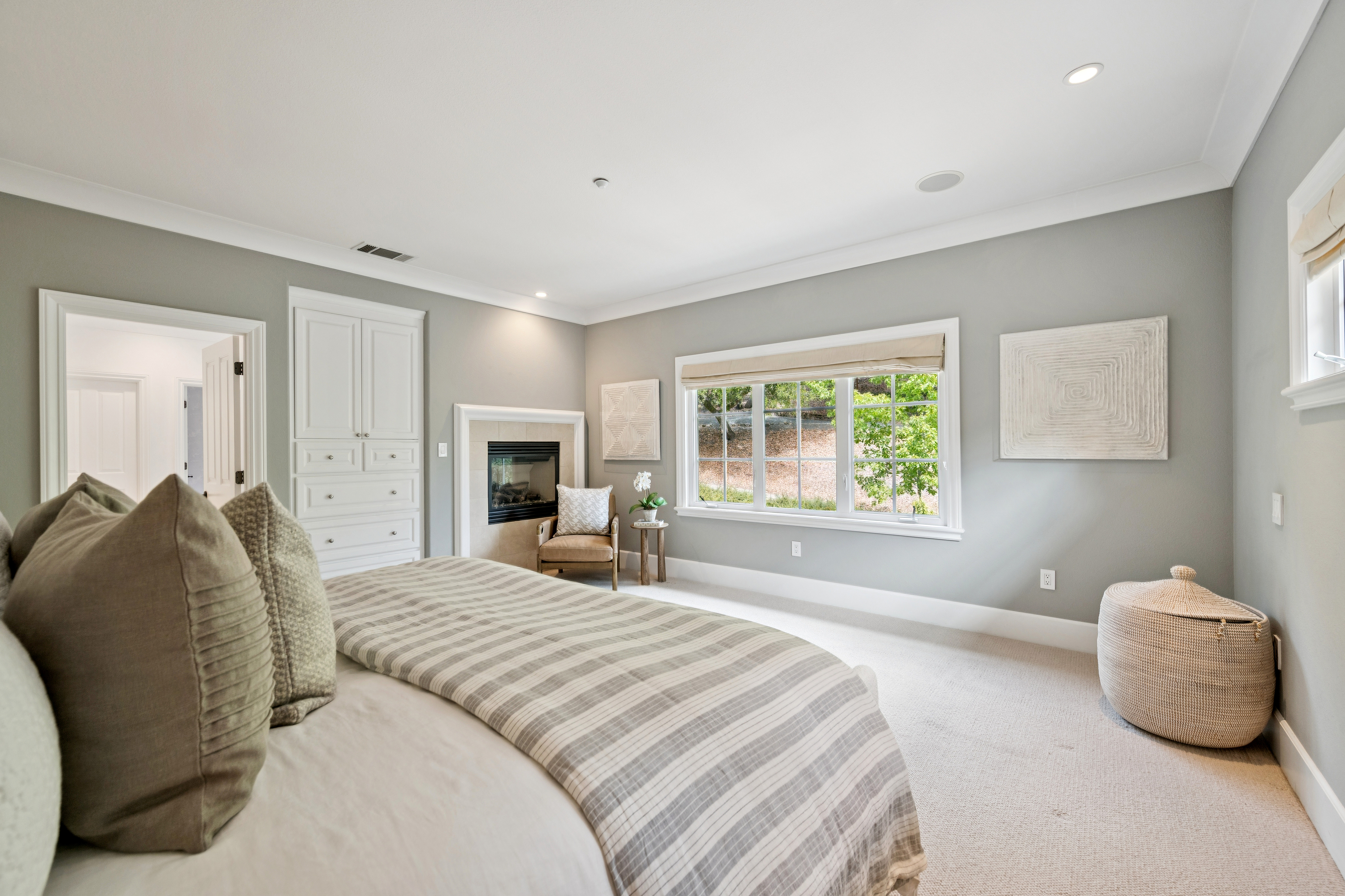 This serene and spacious primary bedroom features soft gray walls, neutral carpeting, and a large window that fills the space with natural light. The room includes a cozy seating area by a built-in fireplace, providing a perfect spot for relaxation. Thoughtful design elements, such as the matching textured wall art and a woven storage basket, add subtle charm and complete the calm, sophisticated aesthetic.