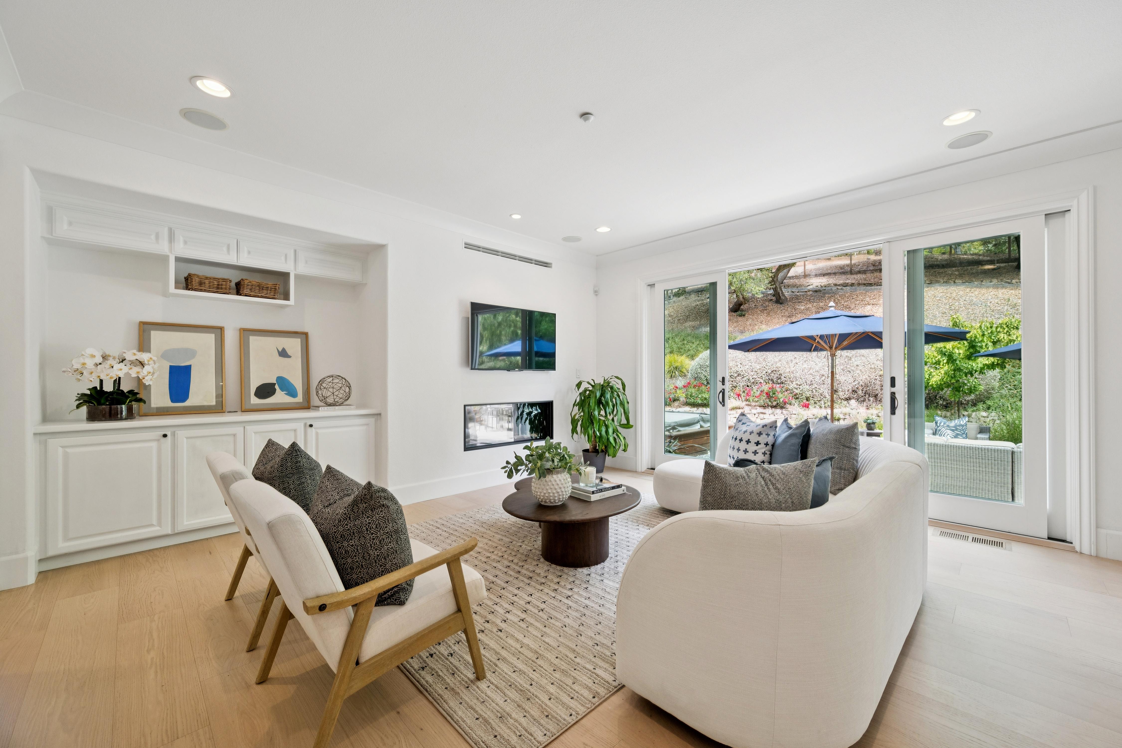 This bright and airy living room features contemporary white cabinetry with open shelving and elegant framed artwork. A curved off-white sofa and matching armchairs are arranged around a modern, dark wood coffee table on a textured area rug. Large floor-to-ceiling sliding glass doors provide a seamless transition to the outdoor patio space, filling the room with natural light.