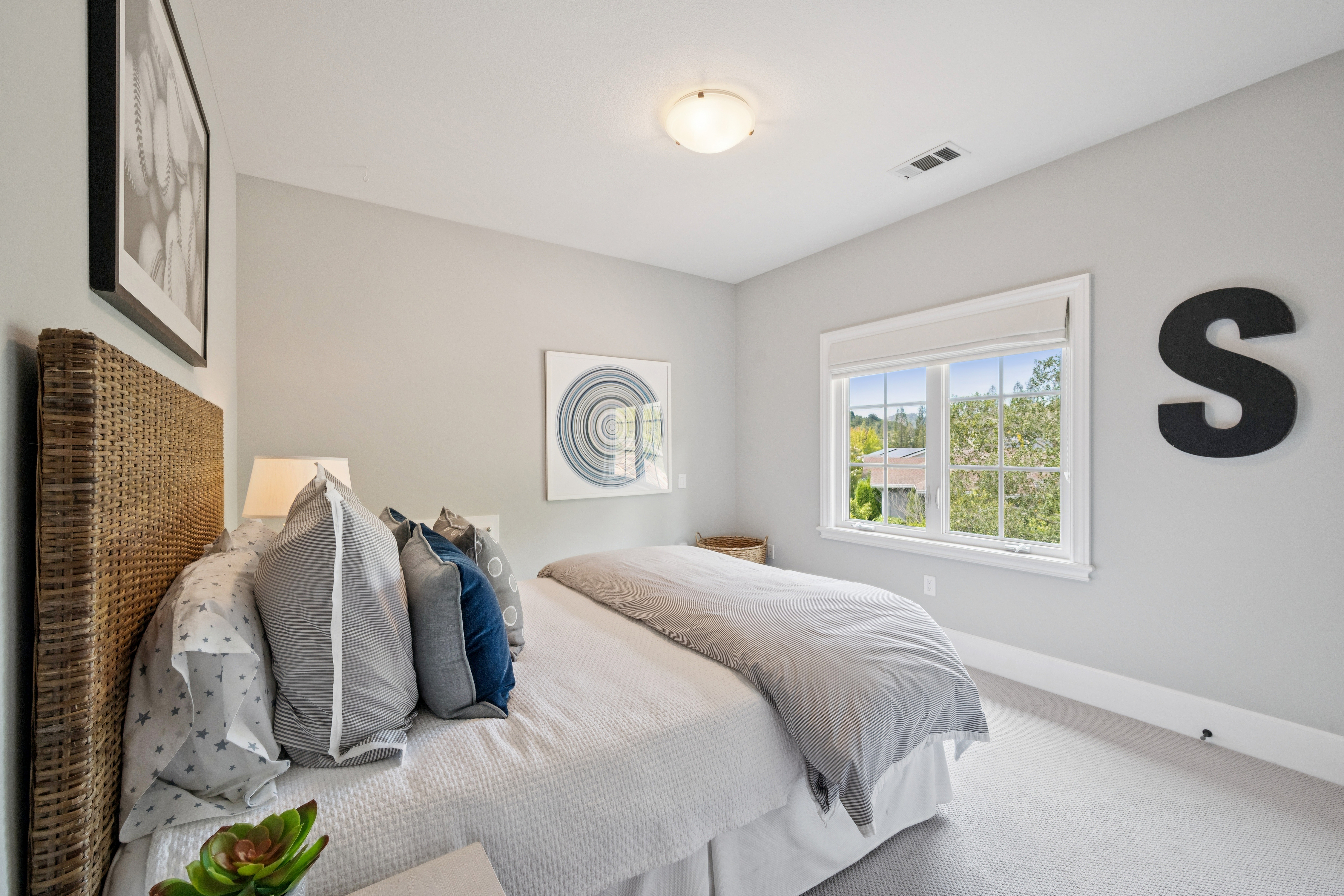This cozy and well-lit bedroom features a textured woven headboard and a layered bed with striped and patterned pillows. A large window provides natural light and a pleasant view, while the walls are decorated with neutral artwork and a decorative 'S' wall accent, creating a clean and inviting atmosphere.