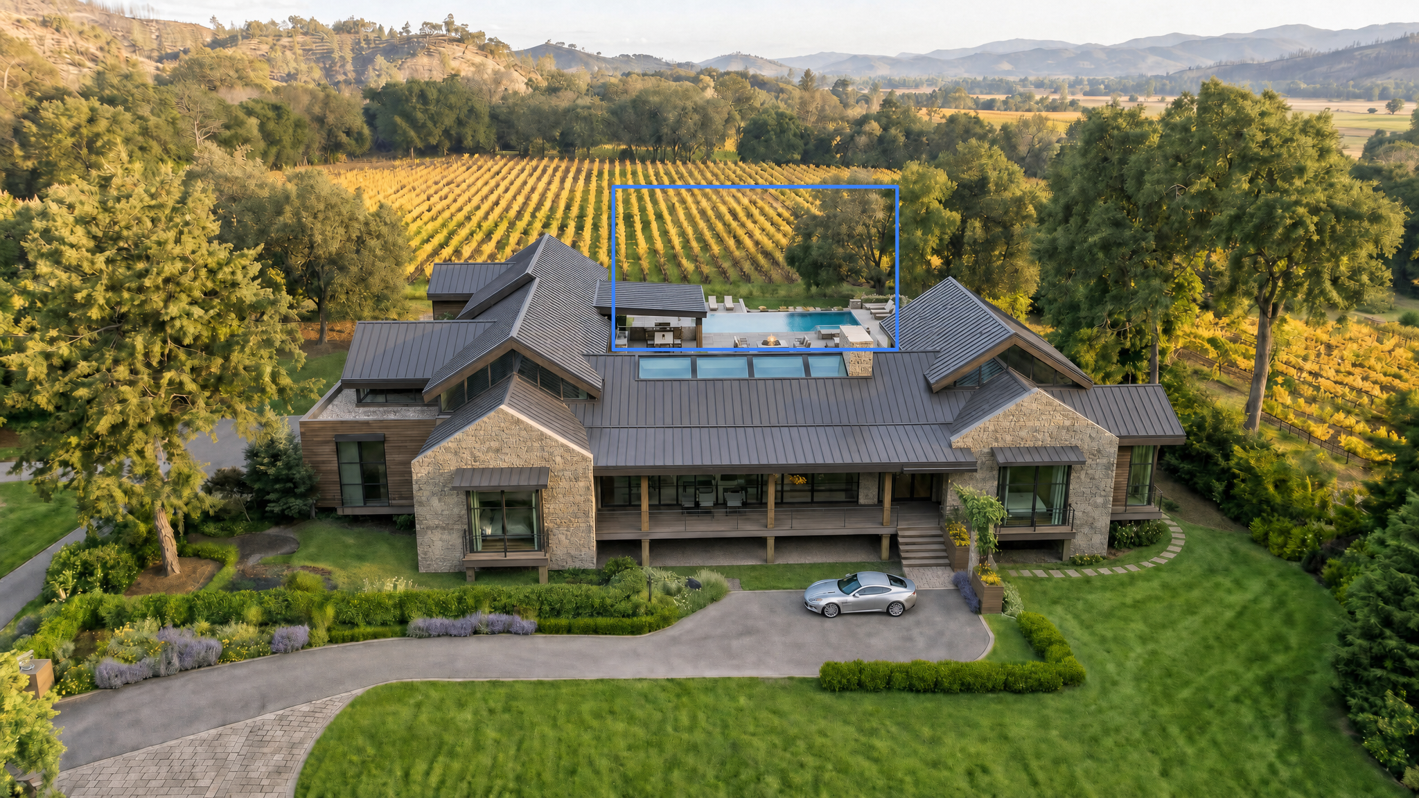 This stunning contemporary estate is captured from an aerial perspective, showcasing its expansive design set against a backdrop of rolling vineyards and distant mountains. Key exterior features include a prominent metal roof, extensive stonework, a dedicated swimming pool area with a fire pit, and a sophisticated landscape design surrounding a private driveway with a luxury vehicle. The home conveys a high-end, elegant aesthetic perfectly integrated into its serene rural surroundings.
