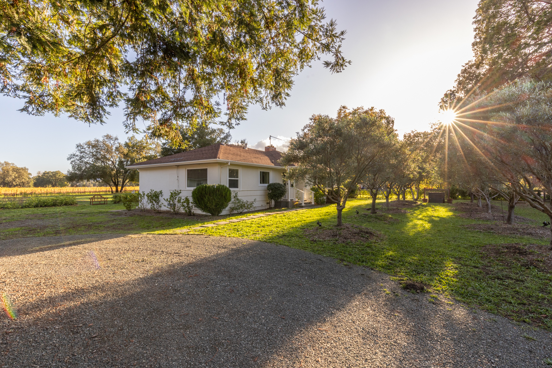 This charming, single-story ranch home is nestled on a spacious, sun-drenched lot. The property features a welcoming gravel driveway, manicured landscaping, and mature trees that frame the white exterior. Its peaceful, pastoral setting conveys a sense of quiet rural living ideal for a residential lifestyle.