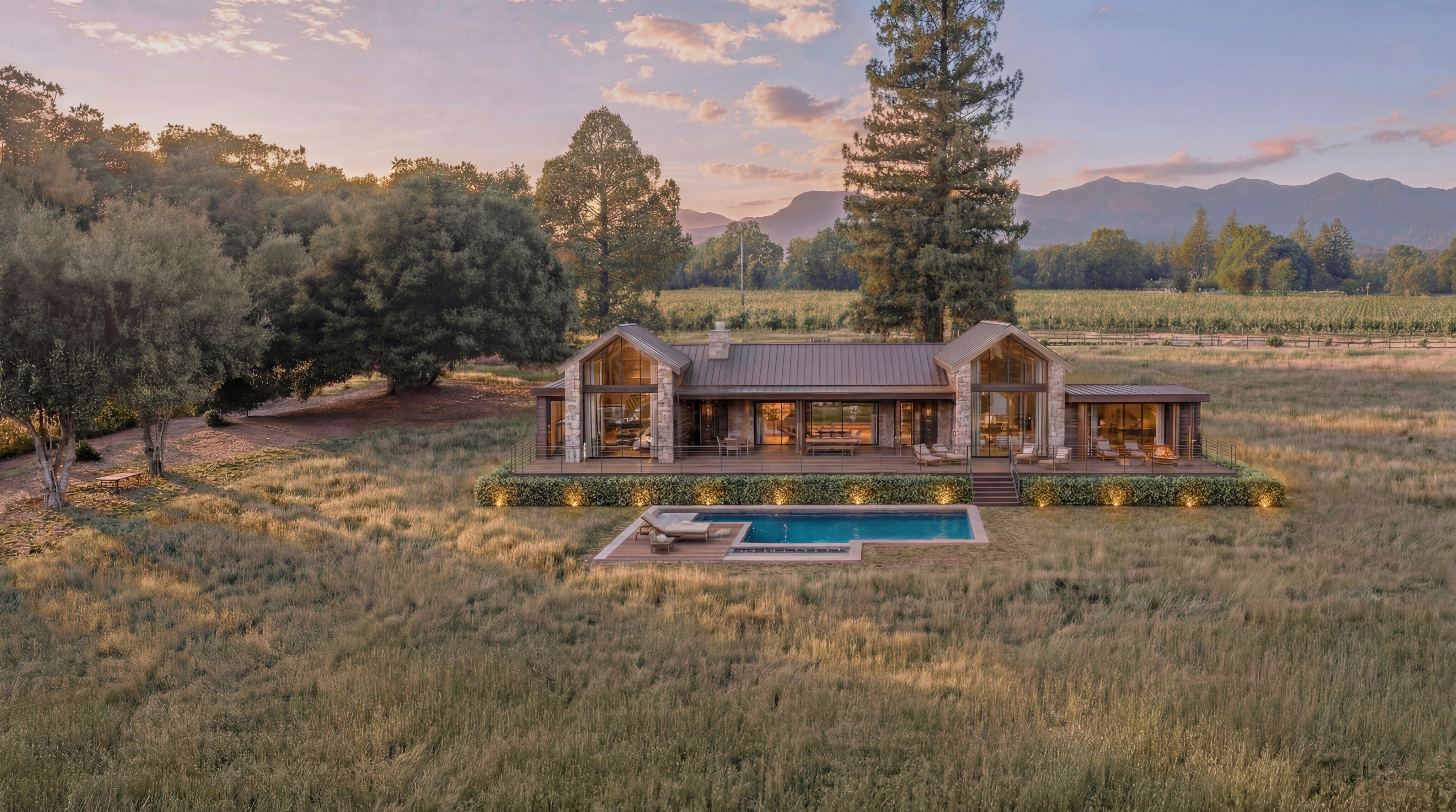 This stunning contemporary farmhouse is captured in an expansive aerial view, nestled within a serene landscape of rolling fields and distant mountains at sunset. The property features a large wooden deck, stone accents, and a tranquil ground-level infinity pool, blending luxury with a rustic, natural environment. With its warm interior lighting and sophisticated design, the residence exudes a luxurious and peaceful atmosphere ideal for modern country living.