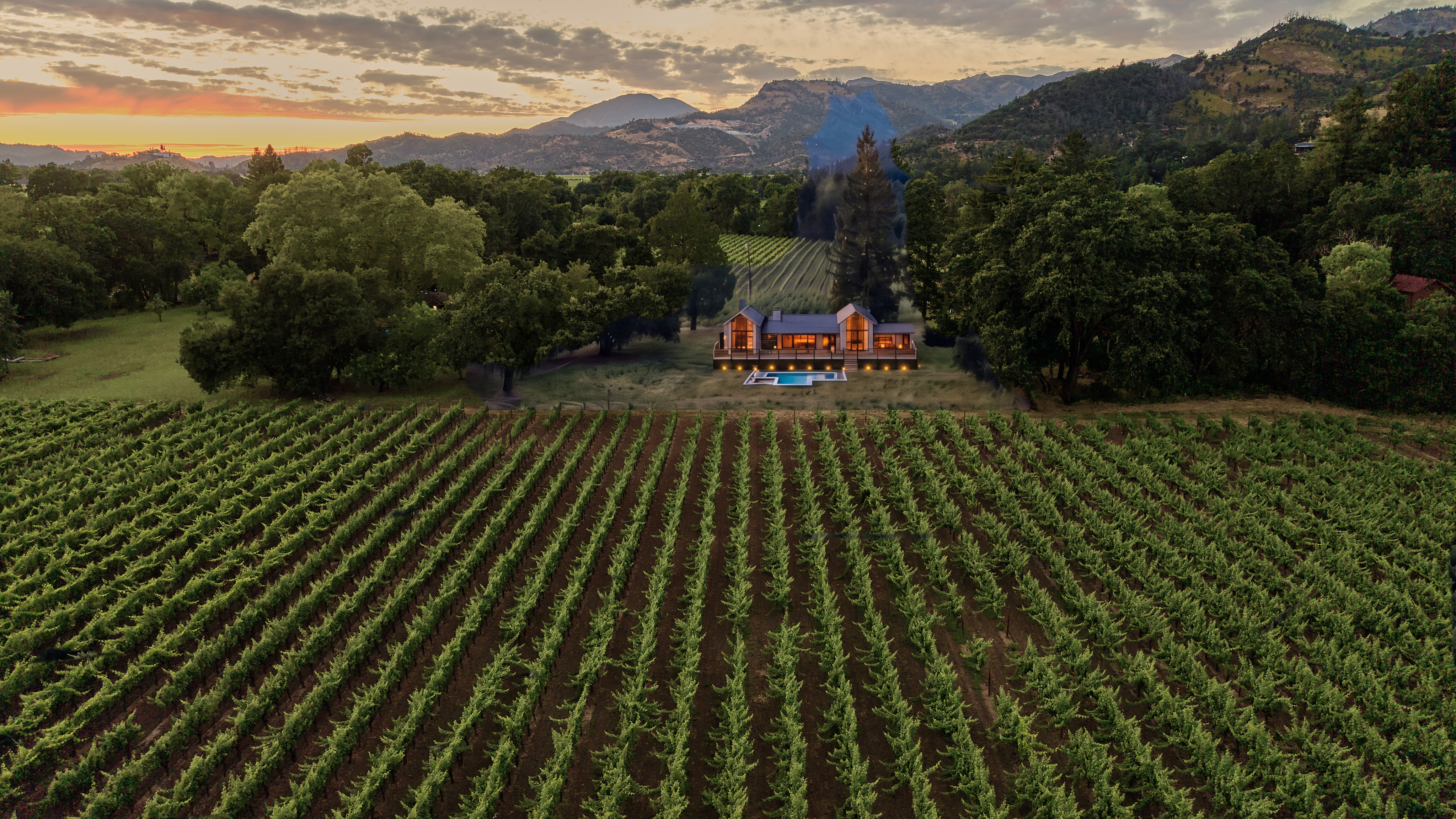 This stunning aerial view captures a contemporary farmhouse-style estate nestled within a lush, expansive vineyard at sunset. The property features a modern residence with warm interior lighting that spills onto a deck overlooking a private pool. Surrounded by mature trees and scenic mountain vistas, the estate offers an atmosphere of secluded luxury and sophisticated rural living.