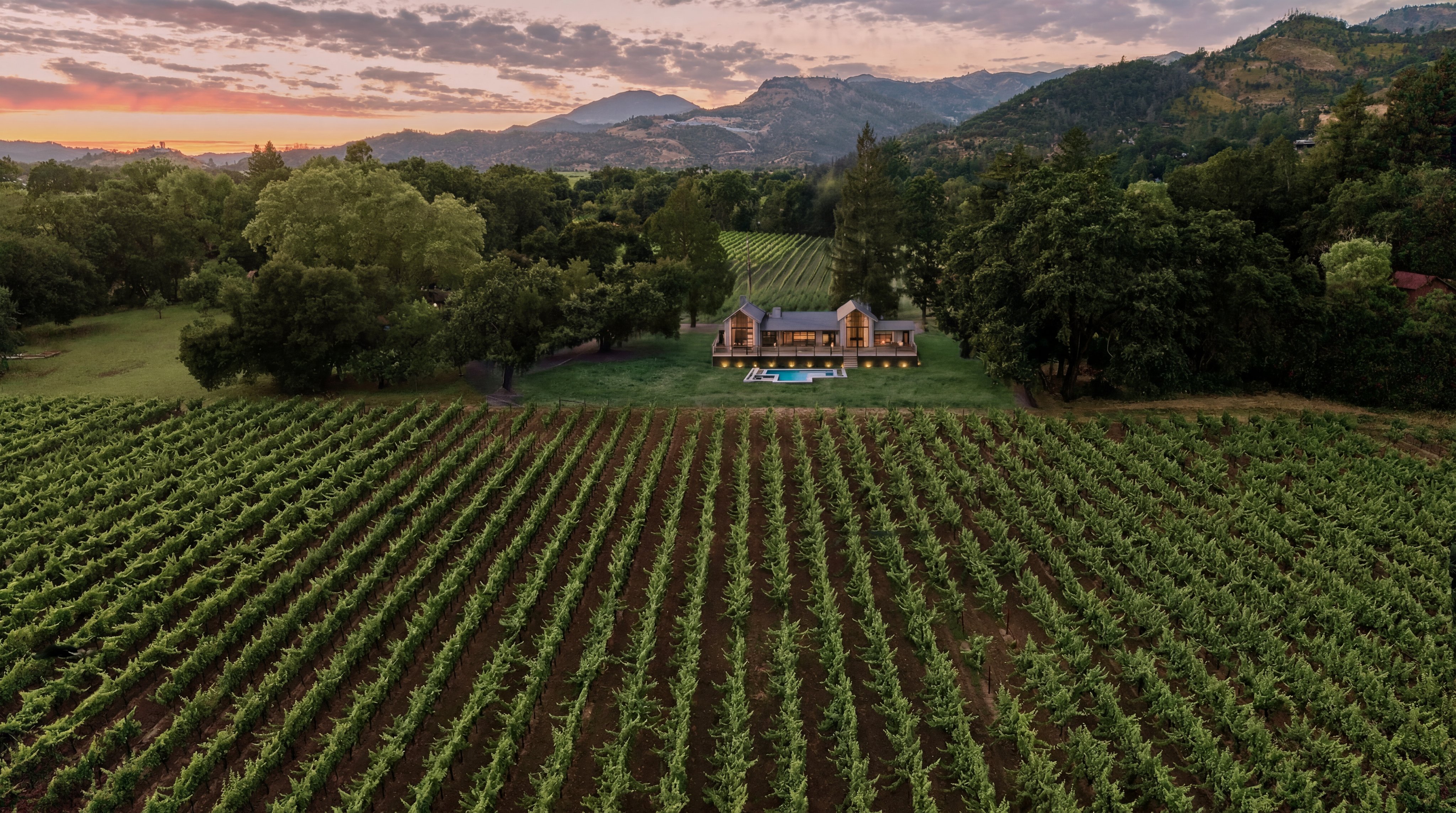 This stunning sunset aerial view captures a luxury residence nestled within a vast vineyard expanse. The contemporary home features peaked rooflines and floor-to-ceiling windows, complemented by a private swimming pool set against a backdrop of lush trees and distant mountains. It offers an evocative atmosphere of tranquility and rural elegance, ideal for those seeking a premium estate property.