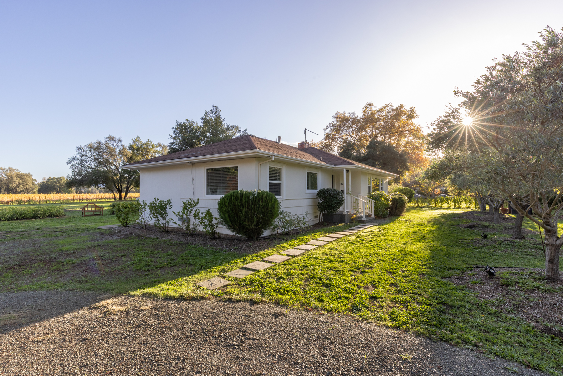 This charming, single-story ranch-style home features a classic white facade and a warm, inviting atmosphere. The property boasts a well-maintained lawn, a stone paver path leading to the entrance, and a sprawling, scenic backdrop including a vineyard. The serene, sun-drenched setting highlights the home's peaceful curb appeal, perfect for anyone looking for a quiet, rural-inspired retreat.