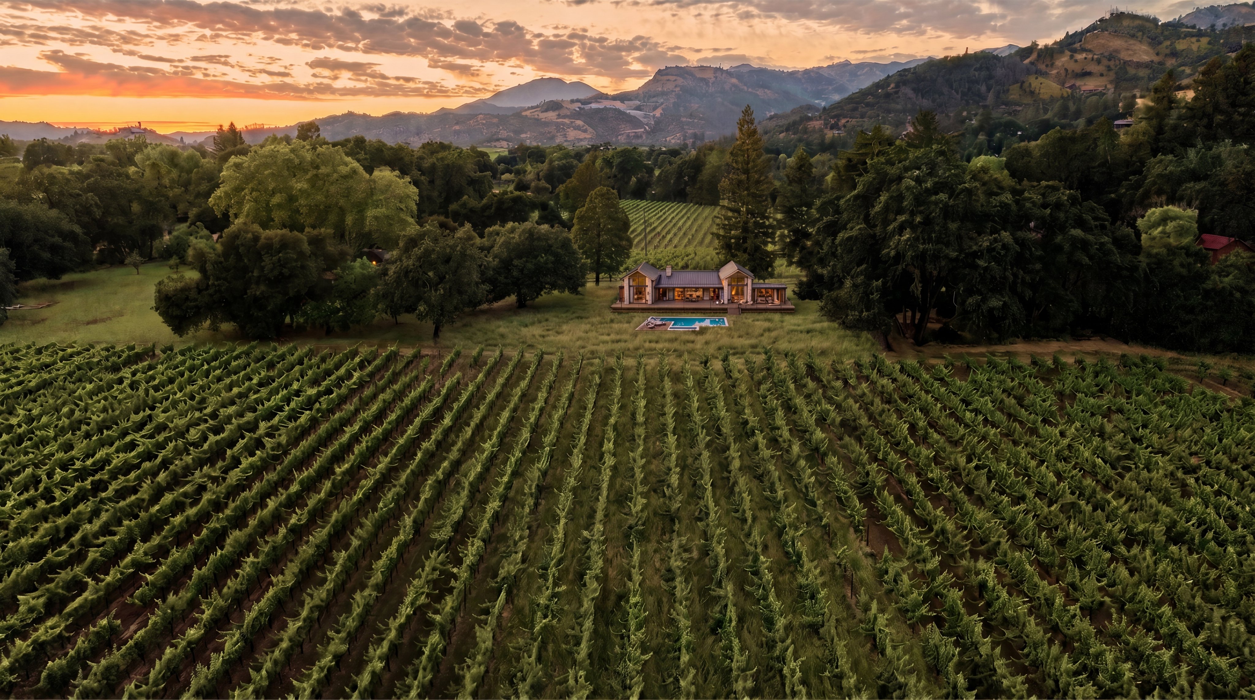 This breathtaking aerial view captures a contemporary farmhouse nestled within a lush, expansive vineyard at sunset. The property features a pristine swimming pool and a wrap-around deck, set against a stunning backdrop of rolling hills and mountains. The serene atmosphere and manicured surroundings emphasize the estate's luxurious and tranquil lifestyle appeal.