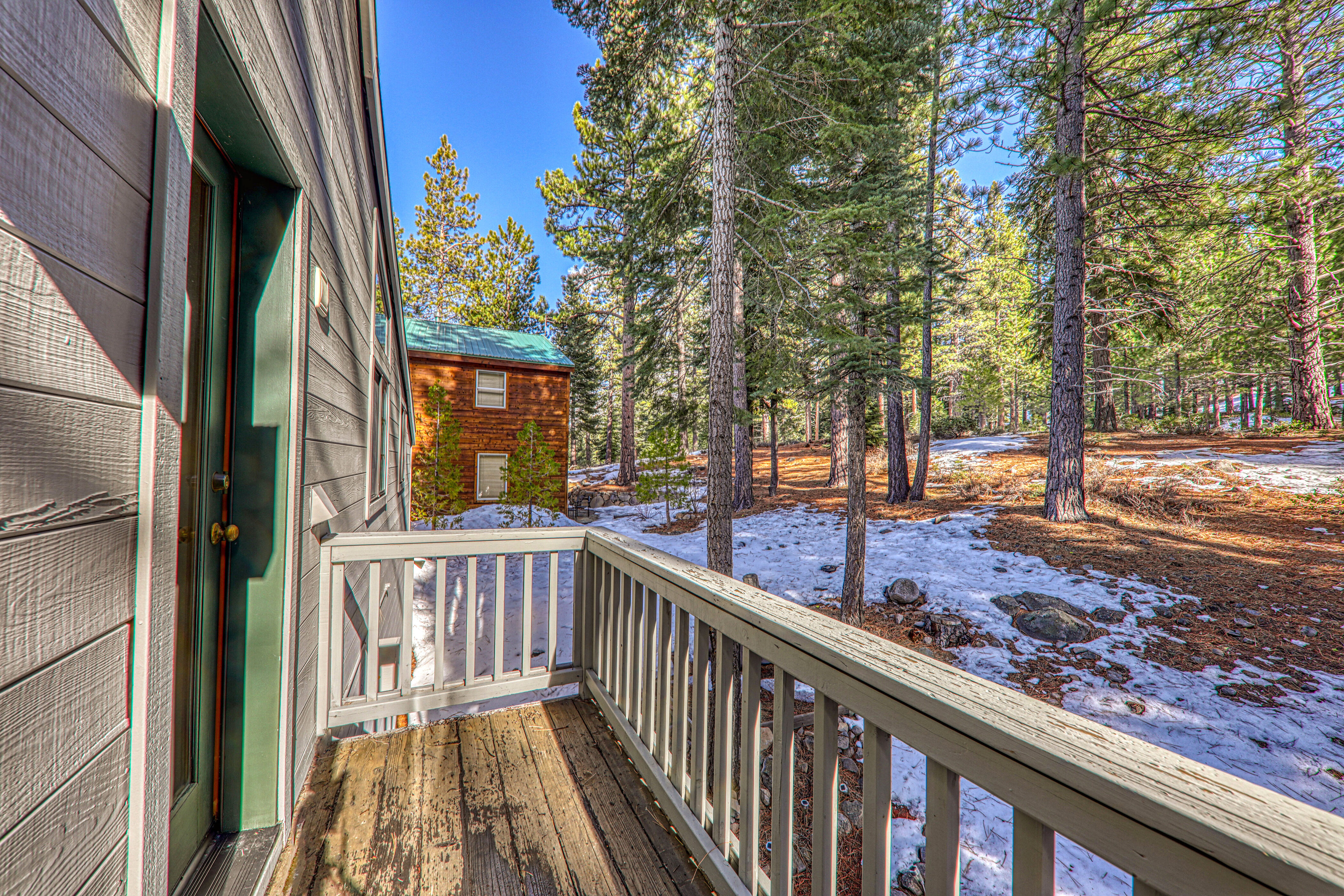 This image shows a wooden deck offering a view of a wooded area with patches of snow. The deck features a simple railing system and provides access to the adjacent structure's entrance, creating a cozy and rustic outdoor living space nestled within a forested setting.