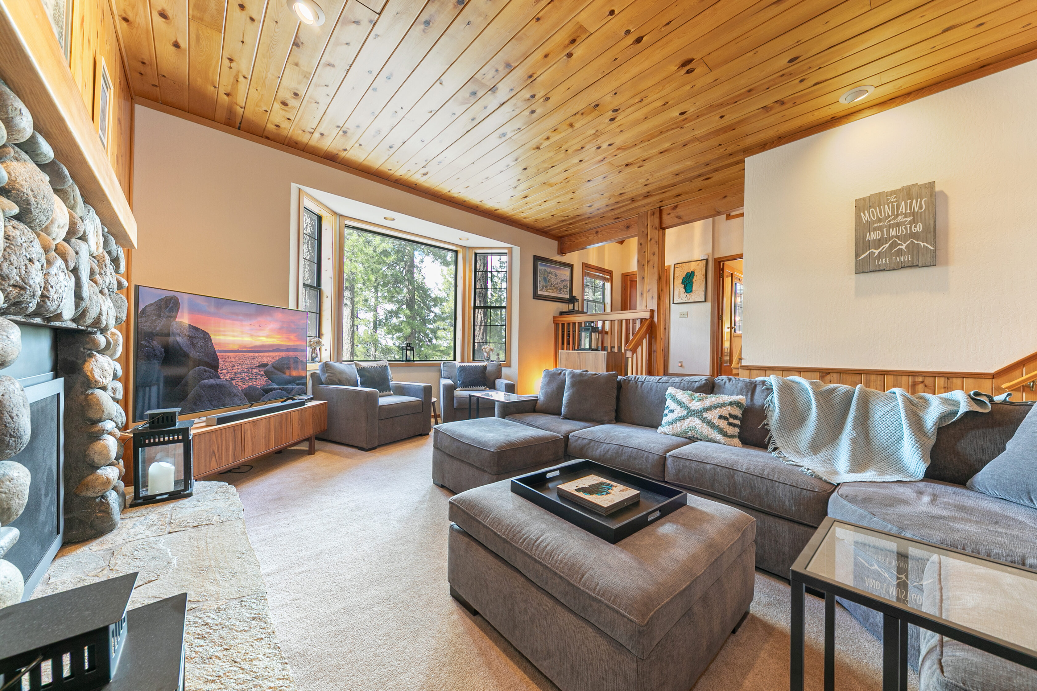 This inviting living room features a rustic cabin aesthetic with a stone fireplace, wood-paneled ceiling, and warm carpeting. A spacious sectional sofa, complemented by a matching ottoman and cozy armchairs, creates an ideal seating area for relaxation. The large bay window allows for abundant natural light and provides serene views of the surrounding forest, reinforcing the home's lodge-like appeal.