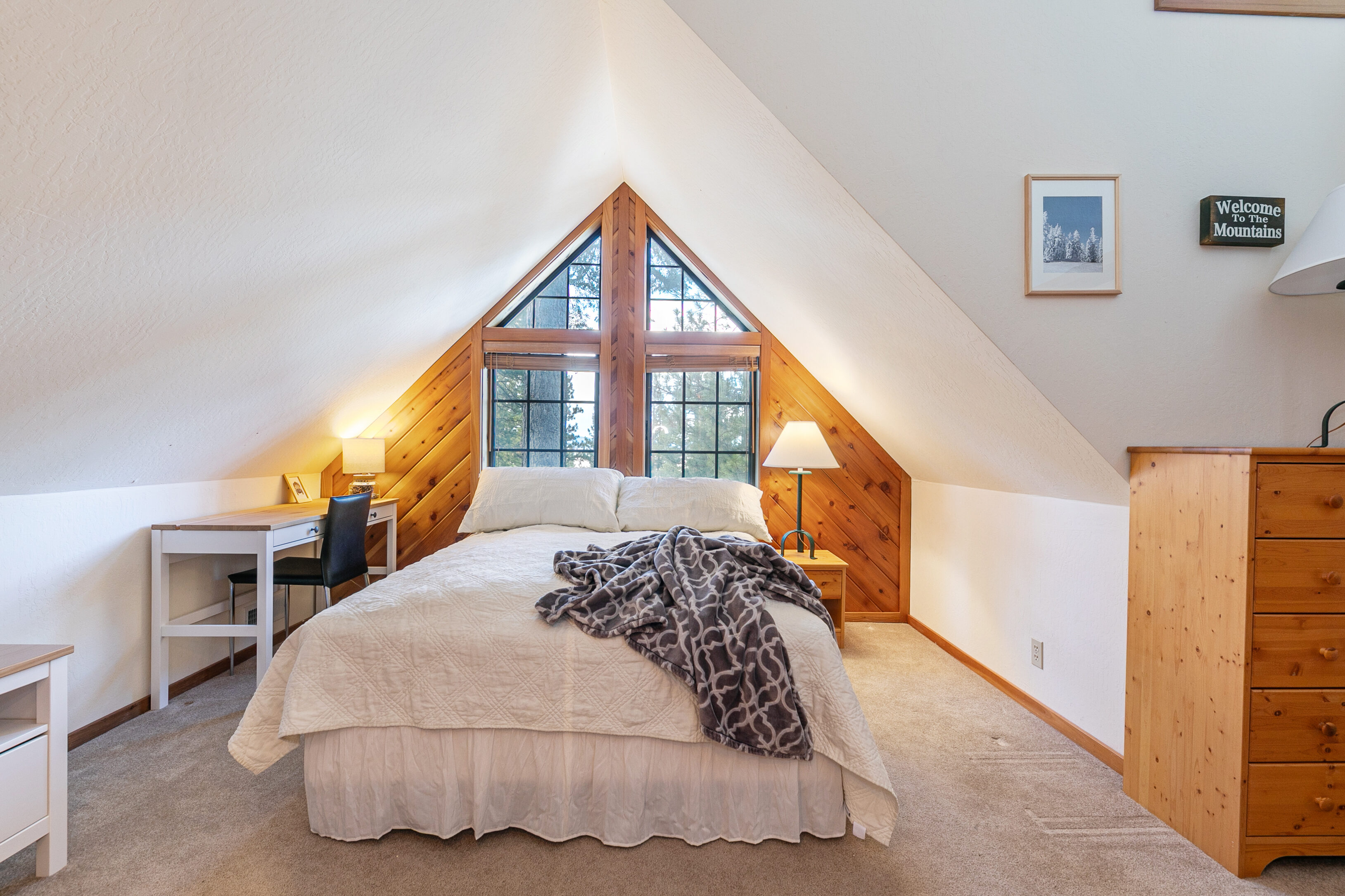 This cozy bedroom is situated in an attic space, featuring a vaulted A-frame ceiling and a large triangular window that brings in natural light. The room is furnished with a comfortable bed, a wooden dresser, and a small desk in the corner, offering a warm and rustic mountain cabin aesthetic. The neutral color palette and functional layout create an inviting and peaceful sleeping area.