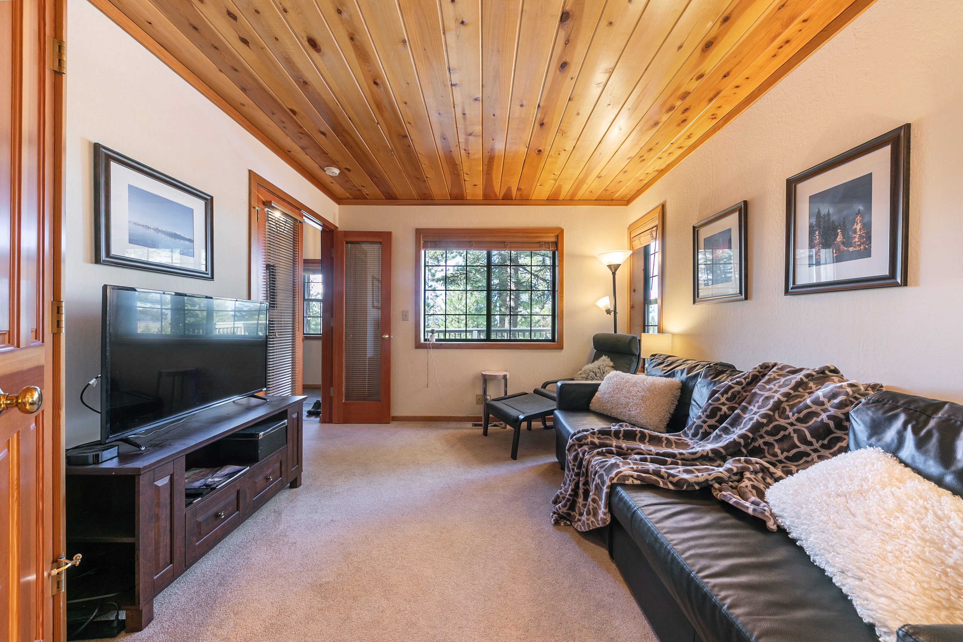 This cozy living room features a warm wood-planked ceiling and comfortable dark leather seating, creating an inviting mountainous retreat feel. The space is well-lit by a combination of natural light from the window and ambient lighting, and it includes a dedicated entertainment area with a large flat-screen TV. With light neutral-toned carpet and tasteful wall art, the room offers a relaxing atmosphere suitable for a mountain or vacation home.