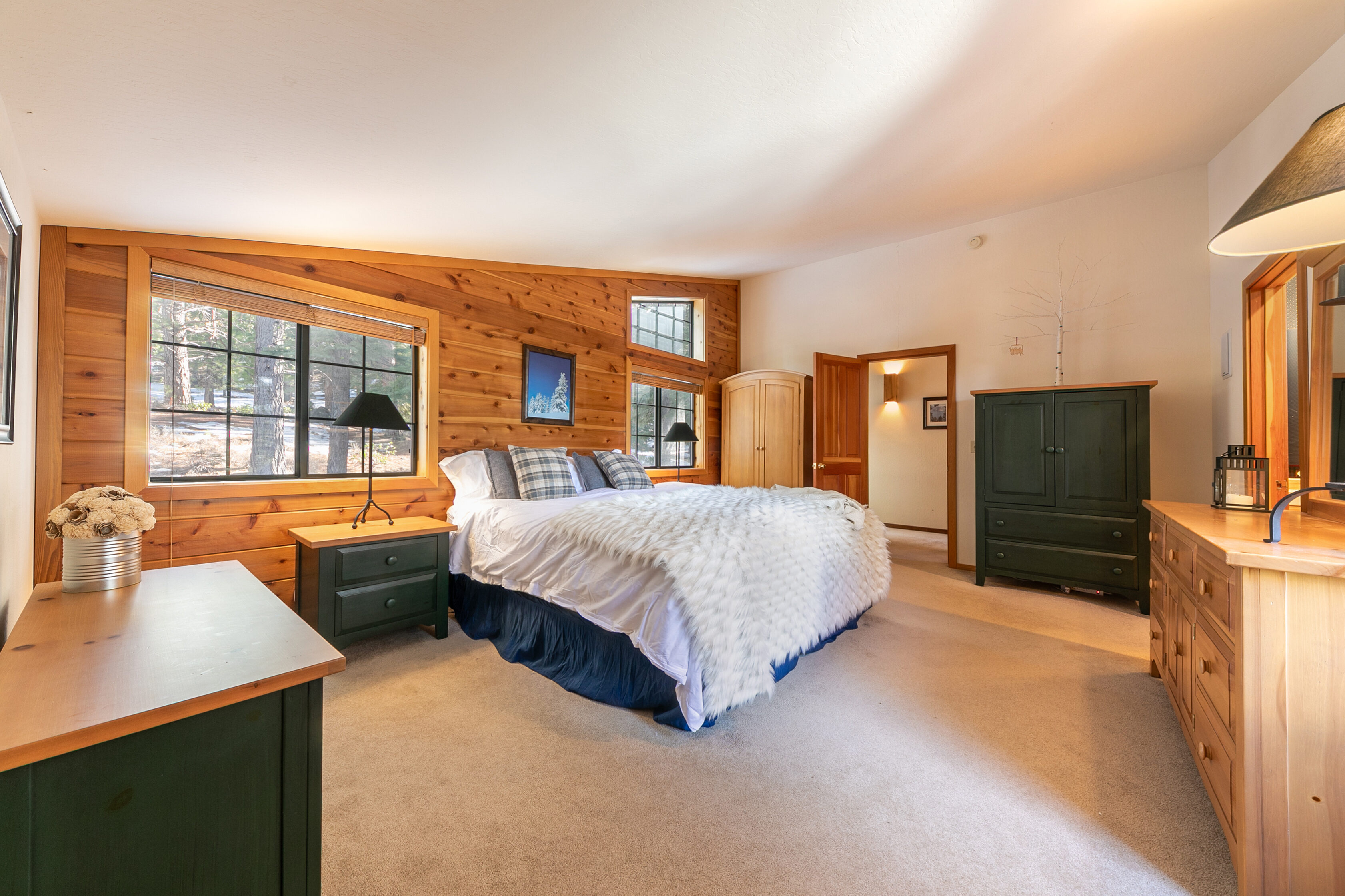 This cozy and rustic primary bedroom features beautiful wood paneling on the main wall and a plush bed accented with a faux-fur throw. The room is well-lit by large windows showing scenery outside and includes matching green-painted nightstands and a large wardrobe. The combination of warm wood tones, comfortable carpeting, and natural light creates an inviting, mountain-style retreat atmosphere.