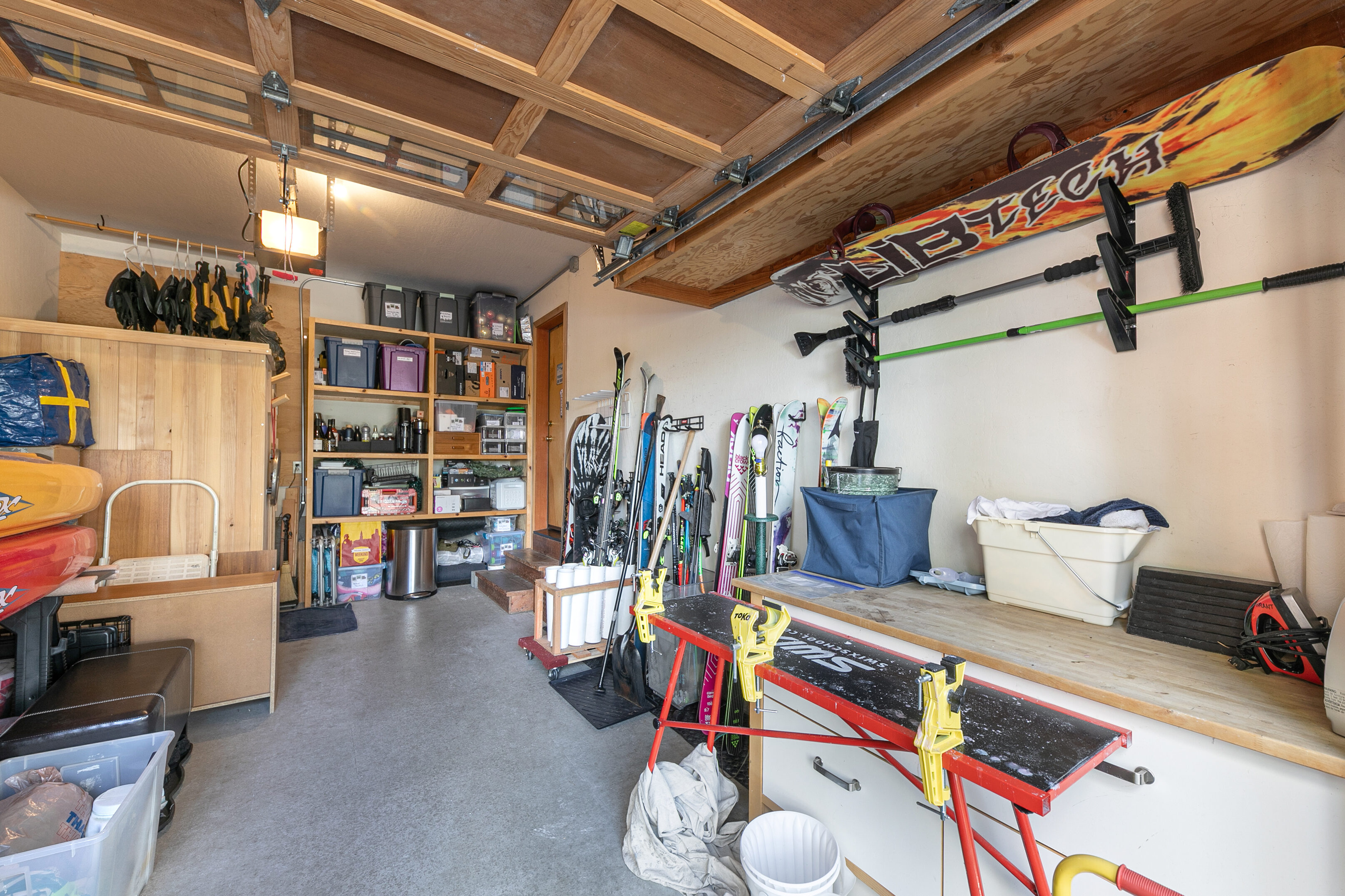 This organized interior garage space serves as a dedicated workshop area for outdoor enthusiasts. It features substantial storage shelving, a workbench equipped with ski-tuning vises, and wall-mounted racks for storing skis, snowboards, and utility items. The space is well-lit and designed for practical functionality, reflecting an active lifestyle storage setup.