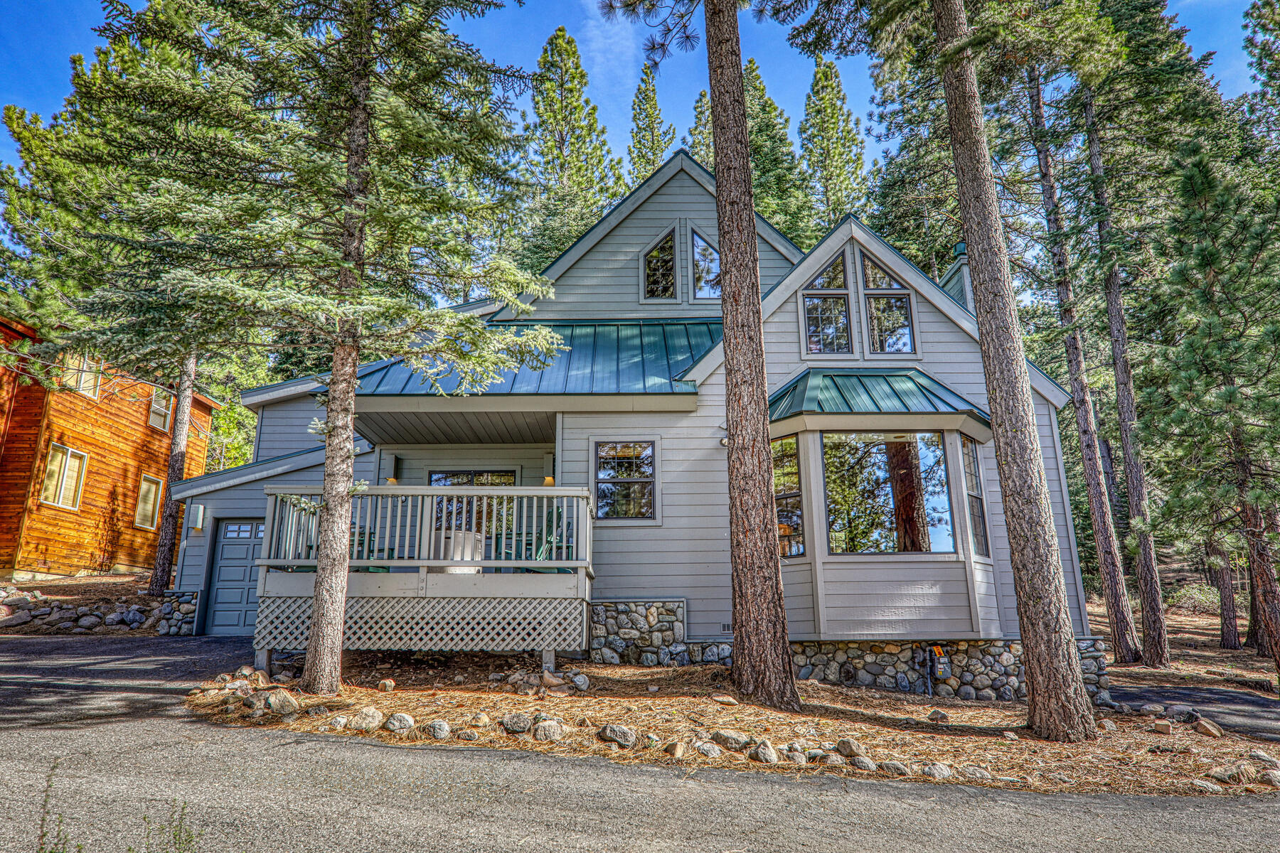 This charming, multi-story mountain home features light grey horizontal siding with a striking green metal roof and a stone foundation. The exterior includes a wooden deck with lattice skirting and a large bay window, surrounded by tall pine trees in a natural forest setting. Its cohesive architectural design offers a classic, rustic retreat appeal perfect for a getaway property.
