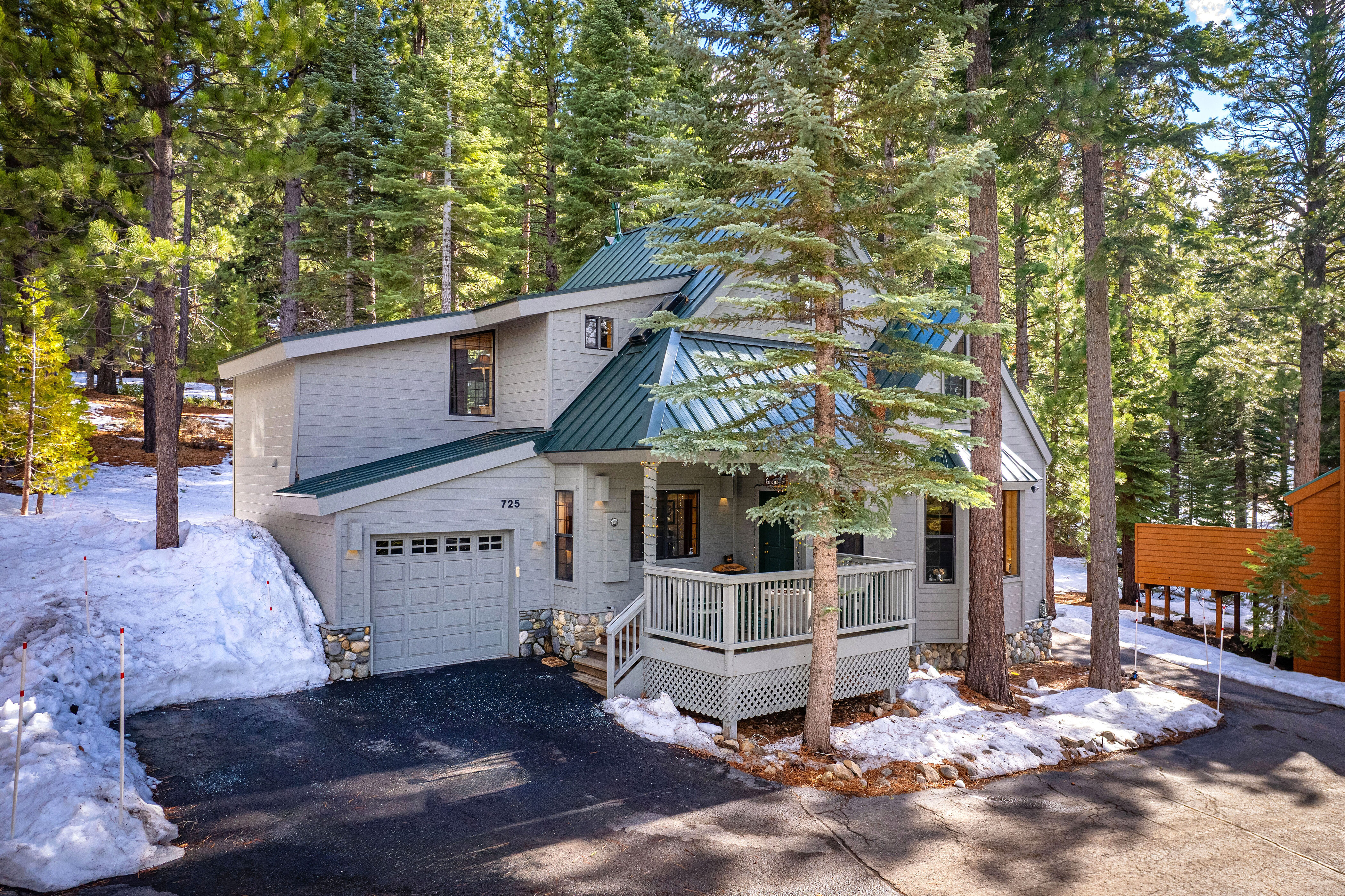 This charming alpine-style home features light gray siding contrasted by a vibrant green metal roof and stone foundation detailing. The property includes an attached garage and a welcoming front deck, all nestled within a wooded, snowy landscape that emphasizes a cozy, mountain-retreat aesthetic.