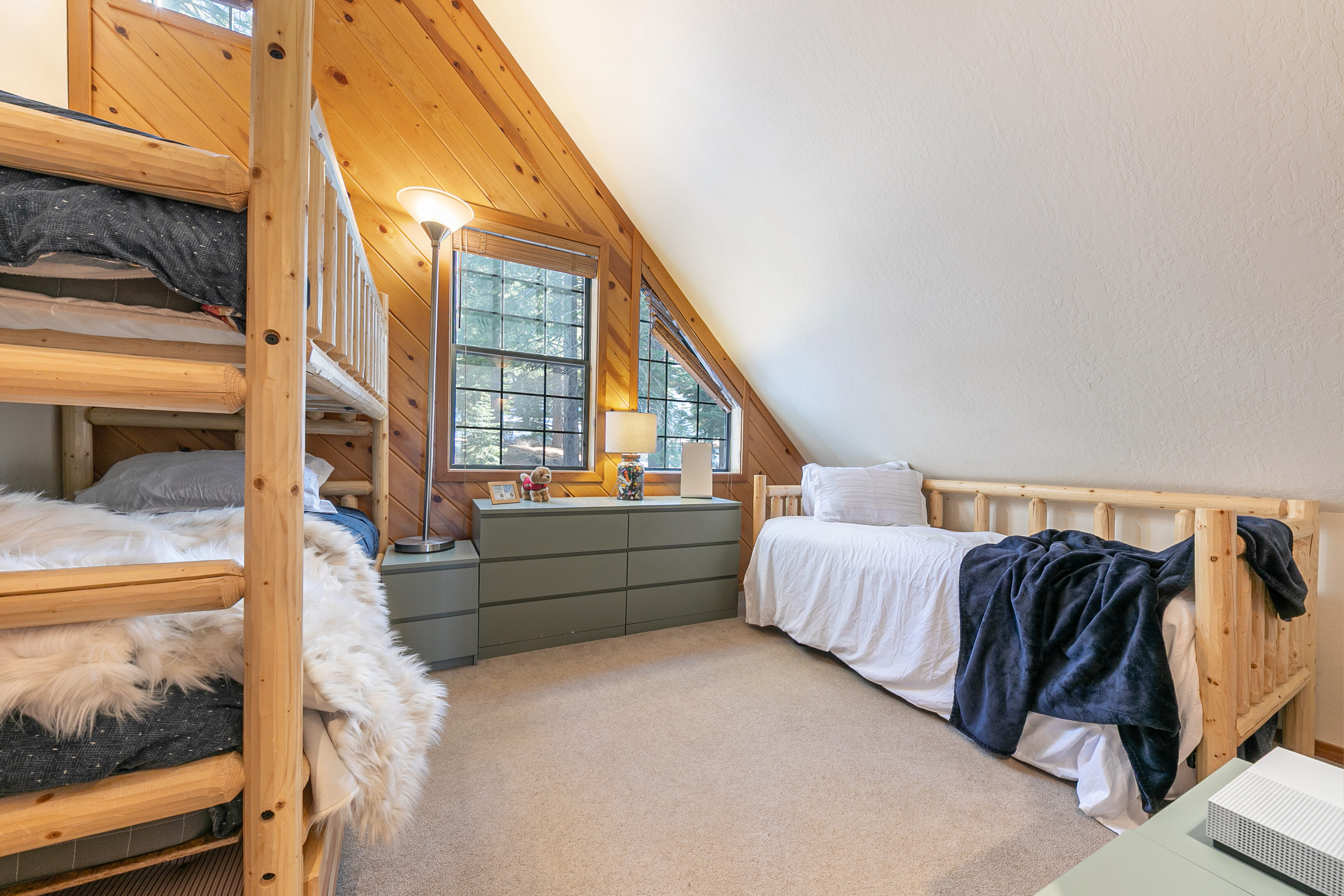 This cozy, rustic-style bedroom features warm wood-paneled walls and a vaulted ceiling, giving it a charming attic-bedroom feel. It is furnished with a wooden bunk bed, a twin bed with a blue throw blanket, and a low dresser. Large windows provide natural light, creating an inviting and comfortable space suitable for guests or children.