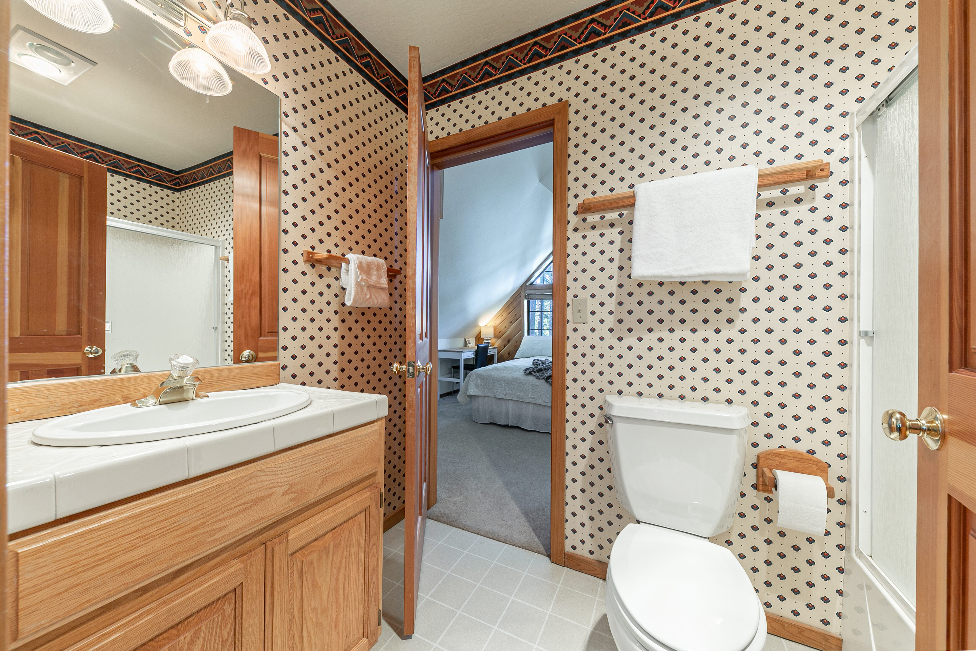 This bathroom features a classic oak vanity with a tiled countertop and a matching wood-framed mirror. The walls are decorated with a neutral, patterned wallpaper and a coordinating decorative border, while a white porcelain toilet sits adjacent to a shower stall. The room provides direct access to an adjoining bedroom, offering a functional and compact layout.