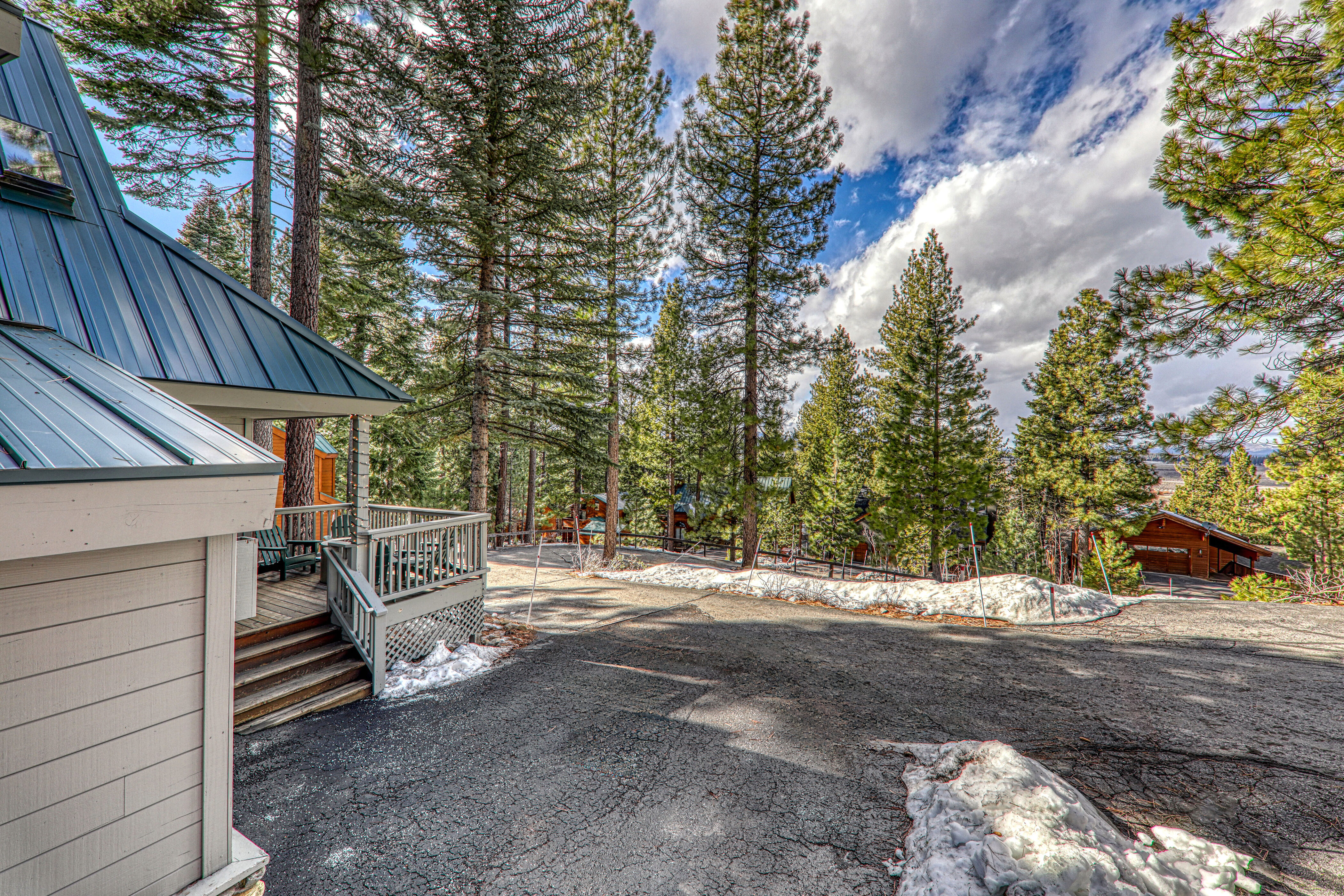 This outdoor view showcases a home's side profile with a focus on an inviting wooden deck equipped with seating. The driveway, partially covered in lingering snow, leads downhill through a scenic, wooded area with mature pine trees under a partially cloudy blue sky. This setting offers a serene, rustic retreat atmosphere characteristic of mountain living.