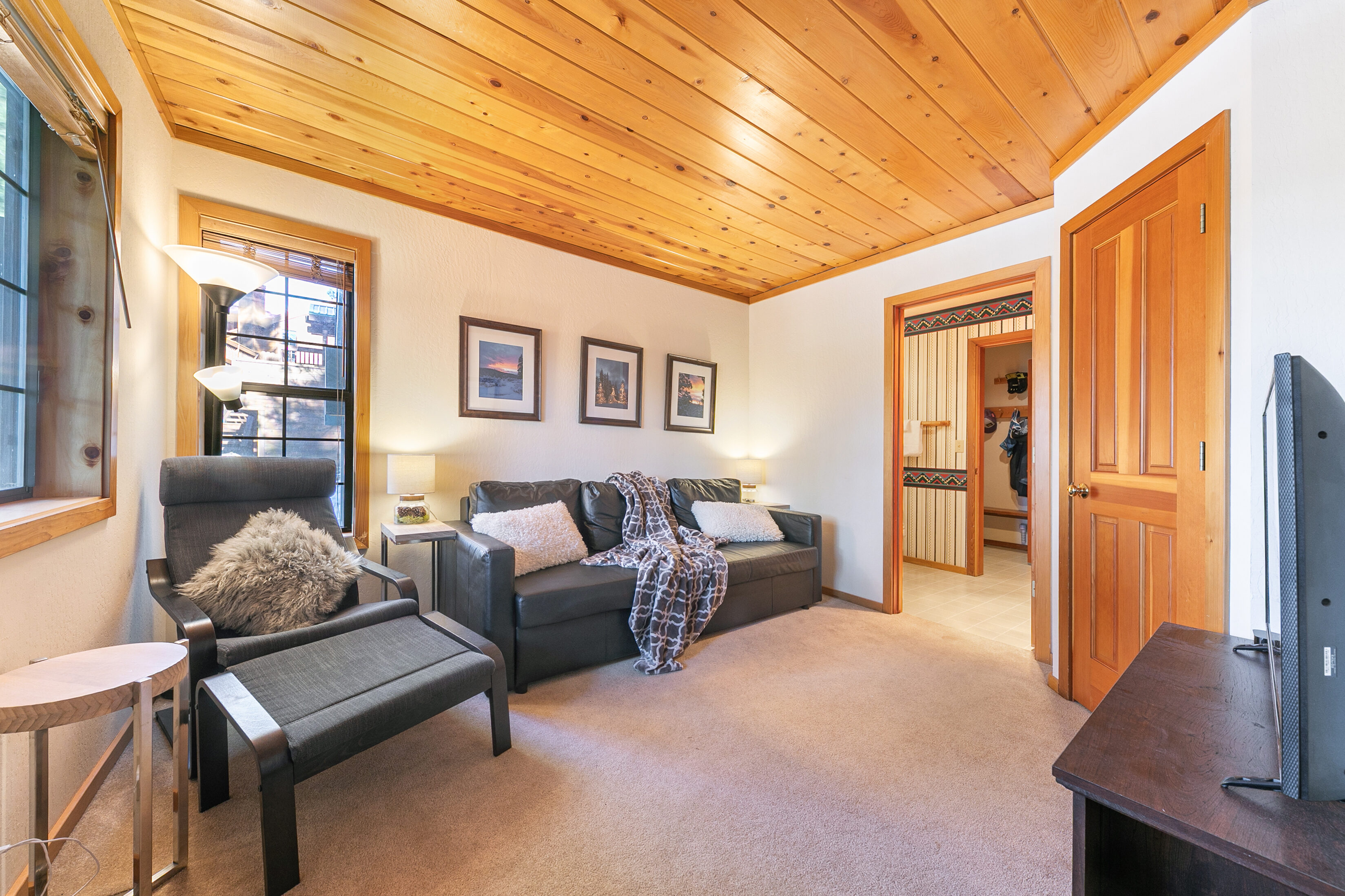 This cozy living room features warm wood-paneled ceilings, a comfortable black leather sofa with a plush throw, and a matching armchair with an ottoman. The space is well-lit with ambient lamps and natural light from the nearby window, accented by framed wall art and carpeted flooring. A doorway leads into a small closet area, giving the room a comfortable and functional feel typical of a mountain retreat or vacation rental.