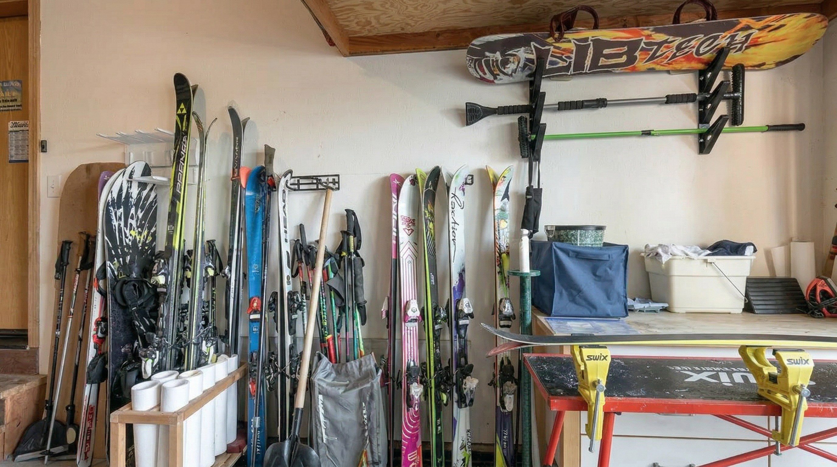 This image displays a well-organized workspace in a garage or utility area, primarily focused on winter sports equipment storage and maintenance. A workbench featuring bright yellow Swix vises is set up for ski tuning, while an extensive collection of skis and a Lib Tech snowboard are stored along the wall. The space is utilitarian and practical, clearly designed to support regular skiing and outdoor activities.