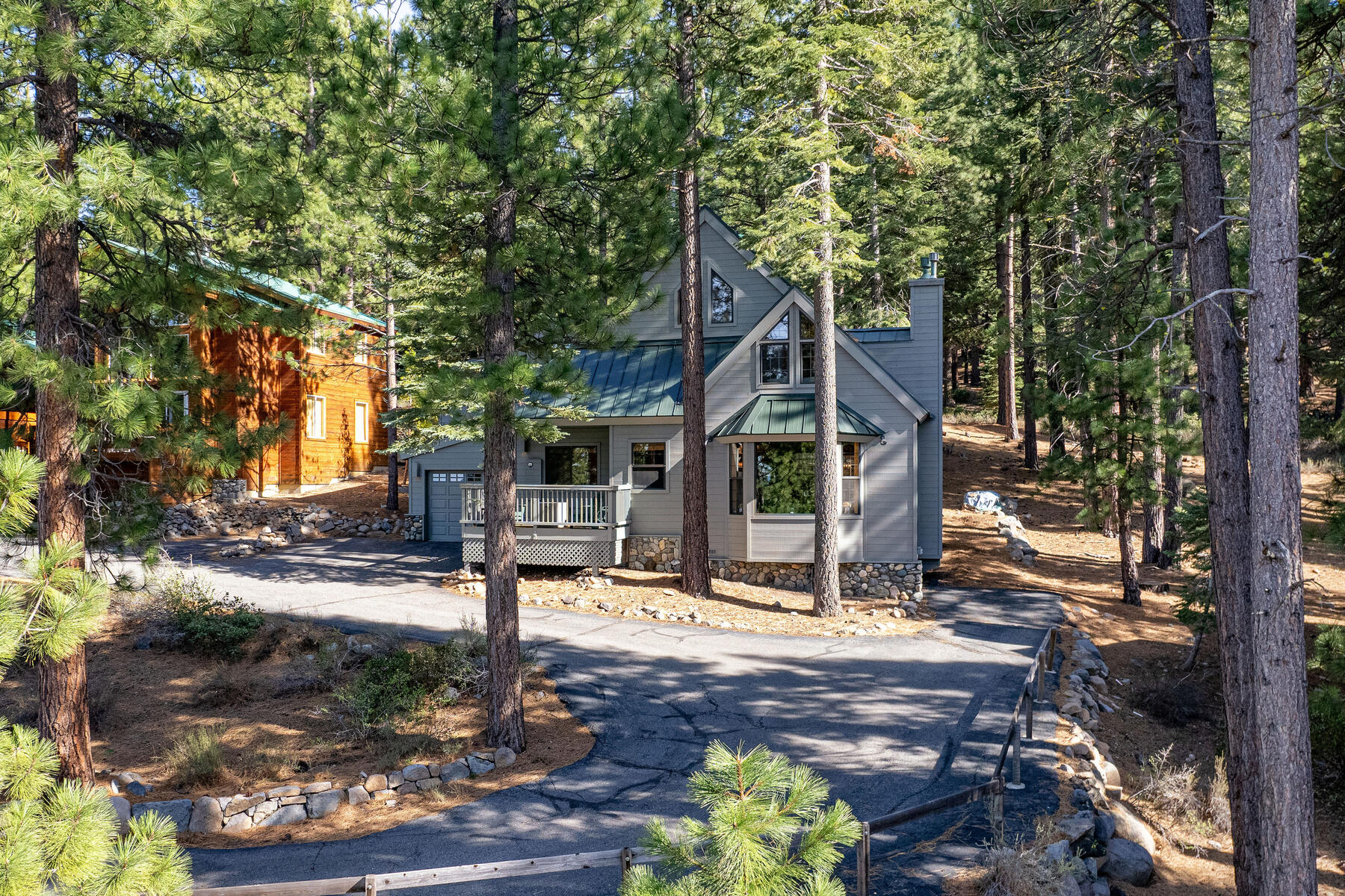 This charming, multi-story home is tucked away in a scenic, pine-forested setting, featuring classic mountain-style architecture with light gray siding and a dark green metal roof. The property includes a paved driveway leading to a built-in garage, a front porch deck, and stone foundation accents that blend harmoniously with the natural landscape. Tucked nicely among mature evergreen trees, the home offers a serene and inviting cabin-like atmosphere.