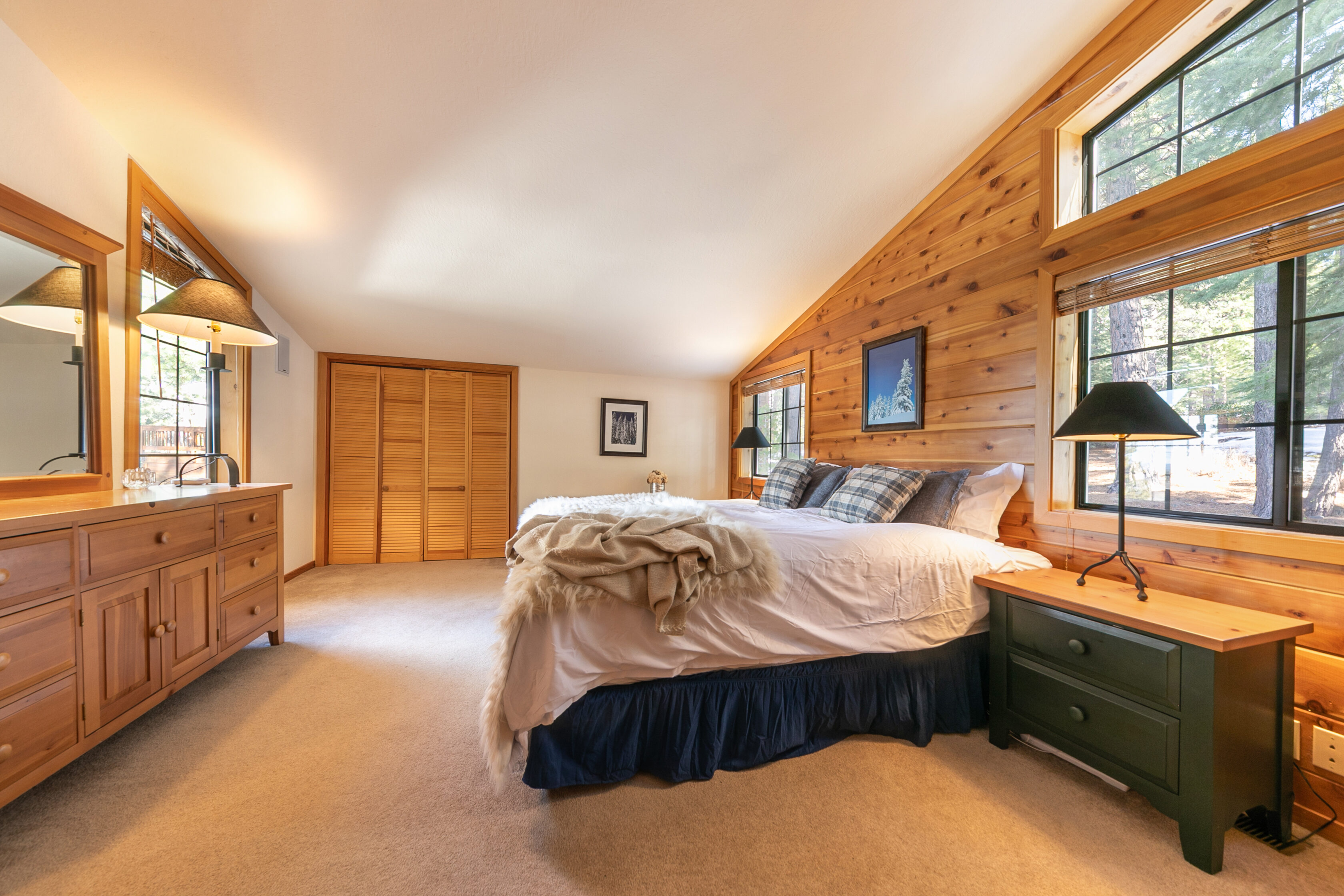 This cozy bedroom features a sloped ceiling and warm wood-paneled walls, creating a charming cabin-style atmosphere. The room is furnished with a comfortable bed with a plush throw, a matching wooden dresser with a mirror, and a dark green nightstand with a lamp. Large windows bring in natural light, offering pleasant views of the surrounding trees.