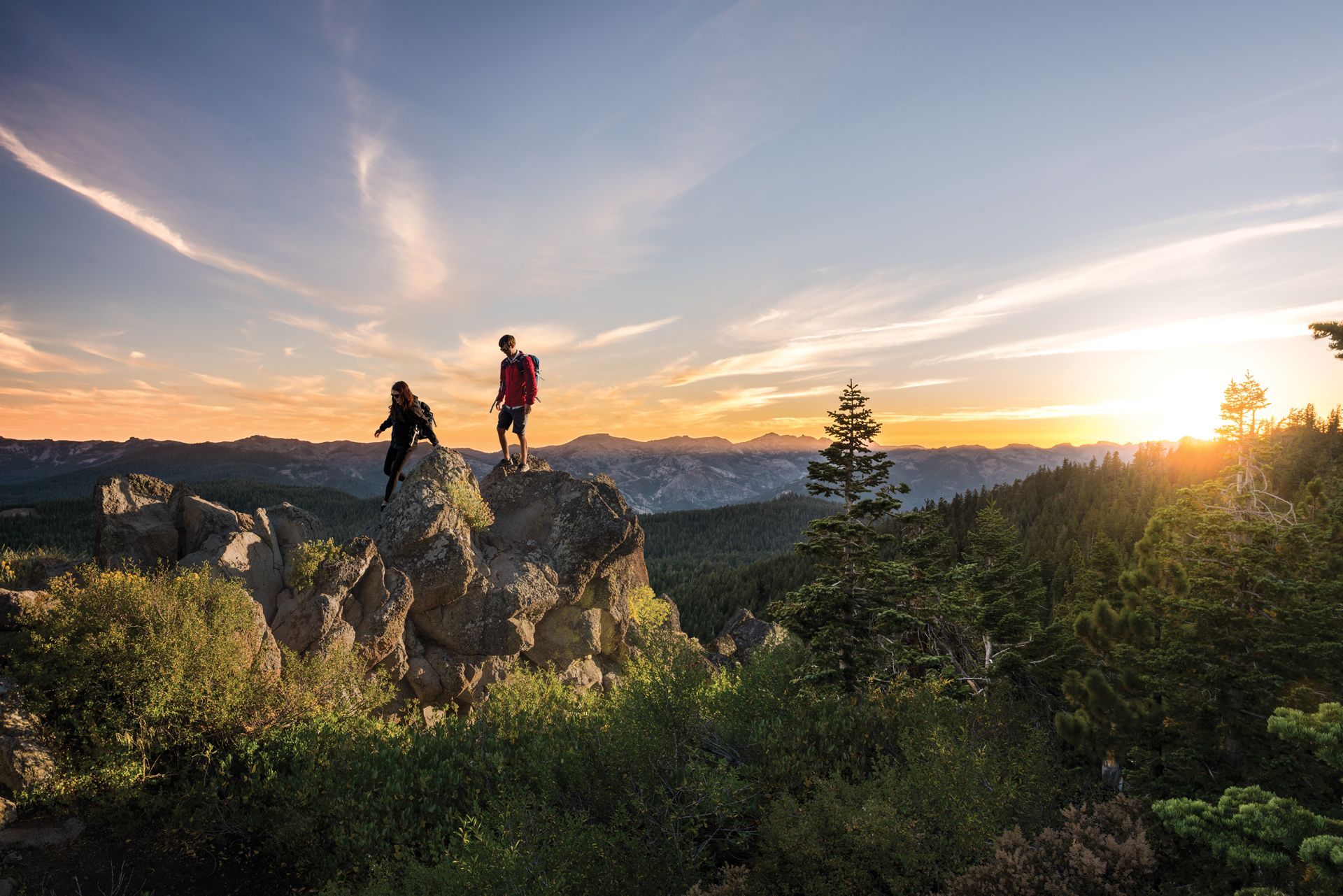 This image features two hikers positioned atop a rugged rock formation, overlooking an expansive mountainous landscape during sunset. The natural terrain includes a dense forest and layered peaks under a vibrant, golden sky. This scenic view highlights the natural beauty and recreational potential associated with the surrounding environment.