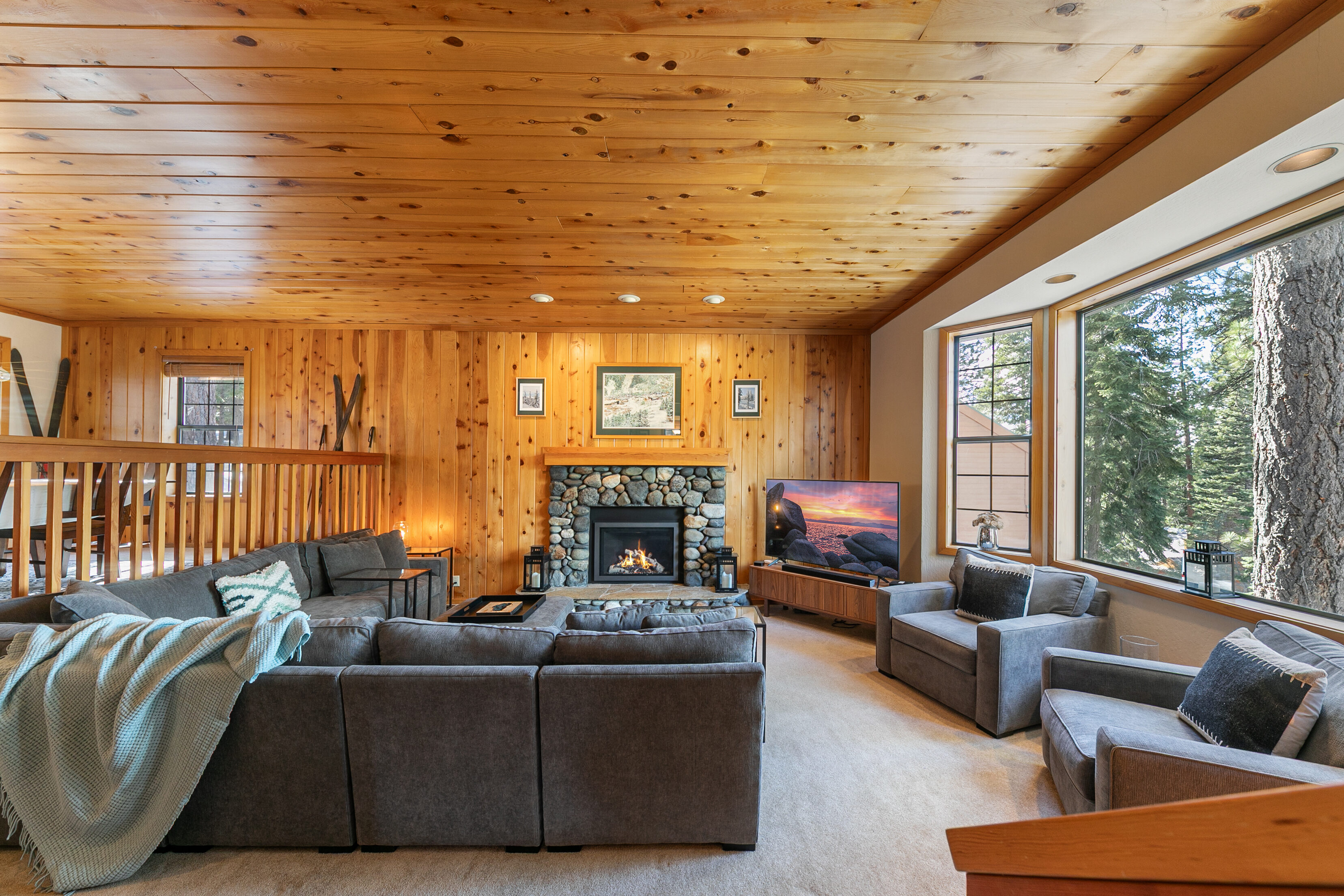 This cozy, rustic living room features warm wood-paneled walls and ceilings, centered around a charming floor-to-ceiling stone fireplace. The room is comfortably furnished with a spacious grey sectional, two armchairs, and floor-to-ceiling windows that offer ample natural light and views of mature trees. Its mountain-lodge aesthetic creates a welcoming and relaxing atmosphere perfect for gathering.
