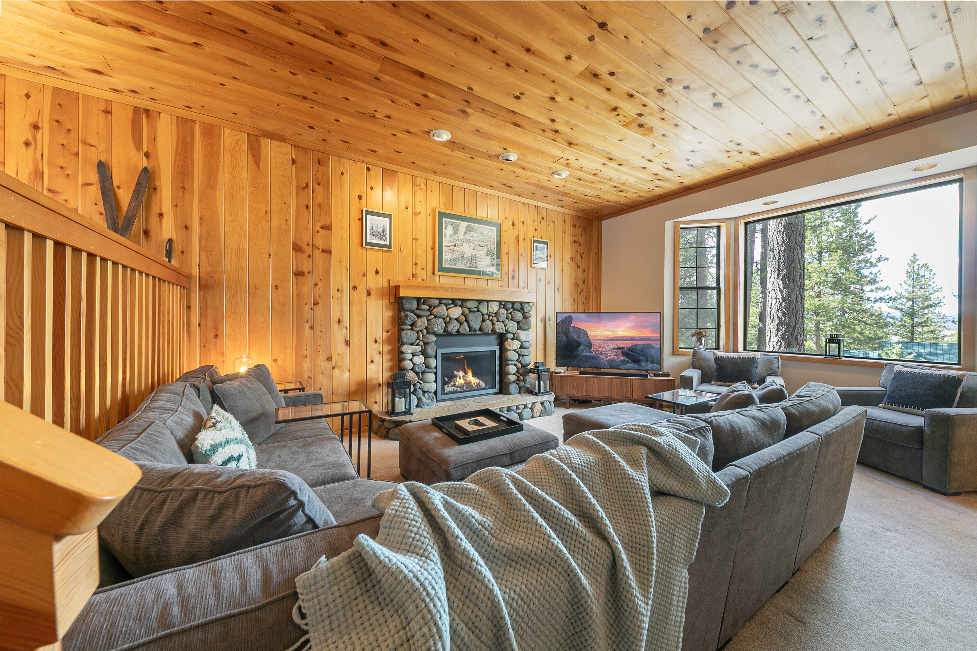 This cozy, rustic-style living room features warm wood-paneled walls and ceilings, creating a classic cabin atmosphere. The room is centered around a striking natural stone fireplace and offers comfortable seating with large gray sectional sofas, making it an ideal space for relaxation. Large windows provide scenic forest views, enhancing the tranquil and inviting nature of the home.