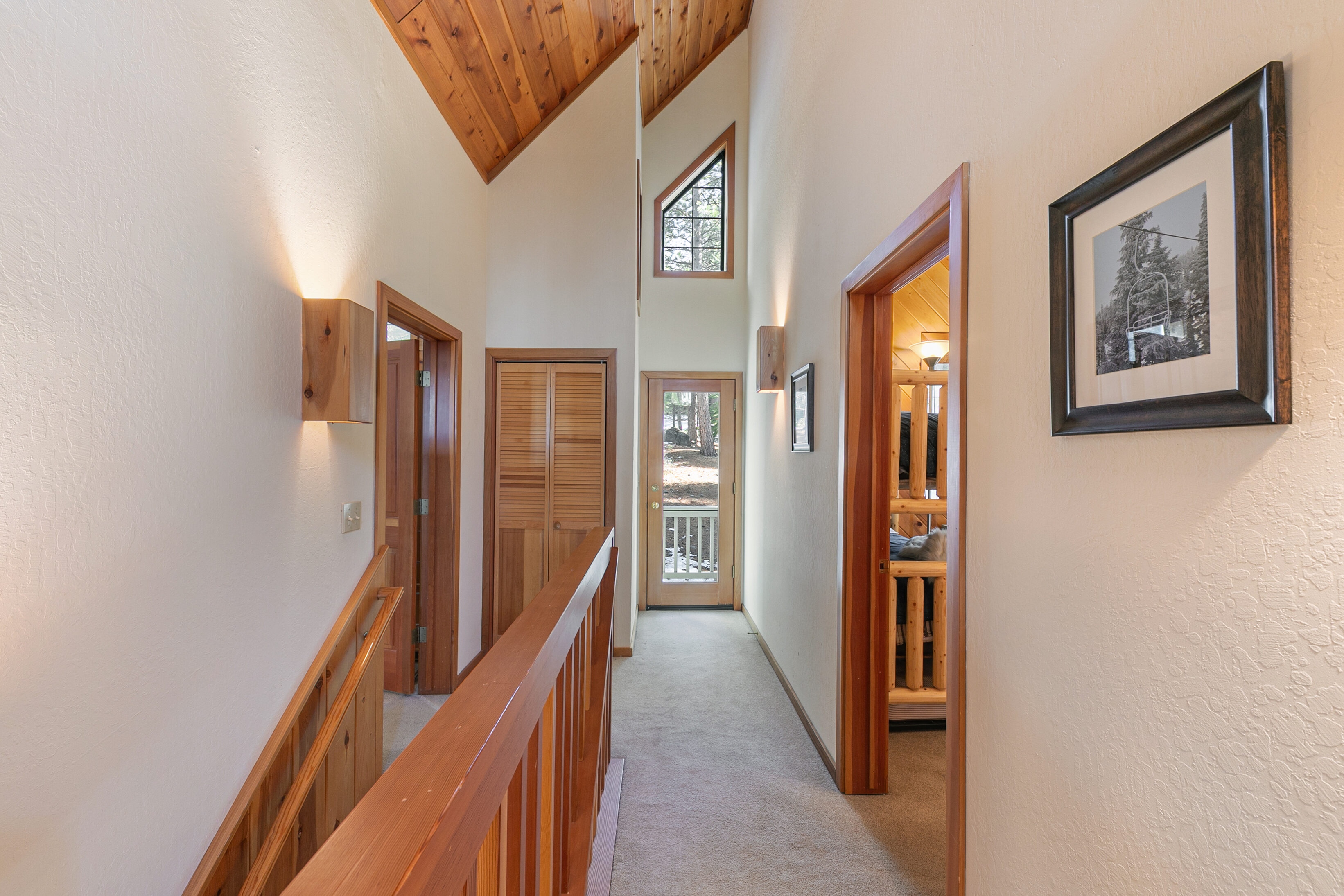 This interior hallway features a vaulted wood-paneled ceiling, beige walls, and carpet flooring, creating a warm, mountain-cabin aesthetic. The space includes a wooden staircase railing, access to multiple rooms, and a doorway leading to an exterior balcony or deck. Architectural interest is added by a unique triangular window and sleek wall-mounted accent lighting.