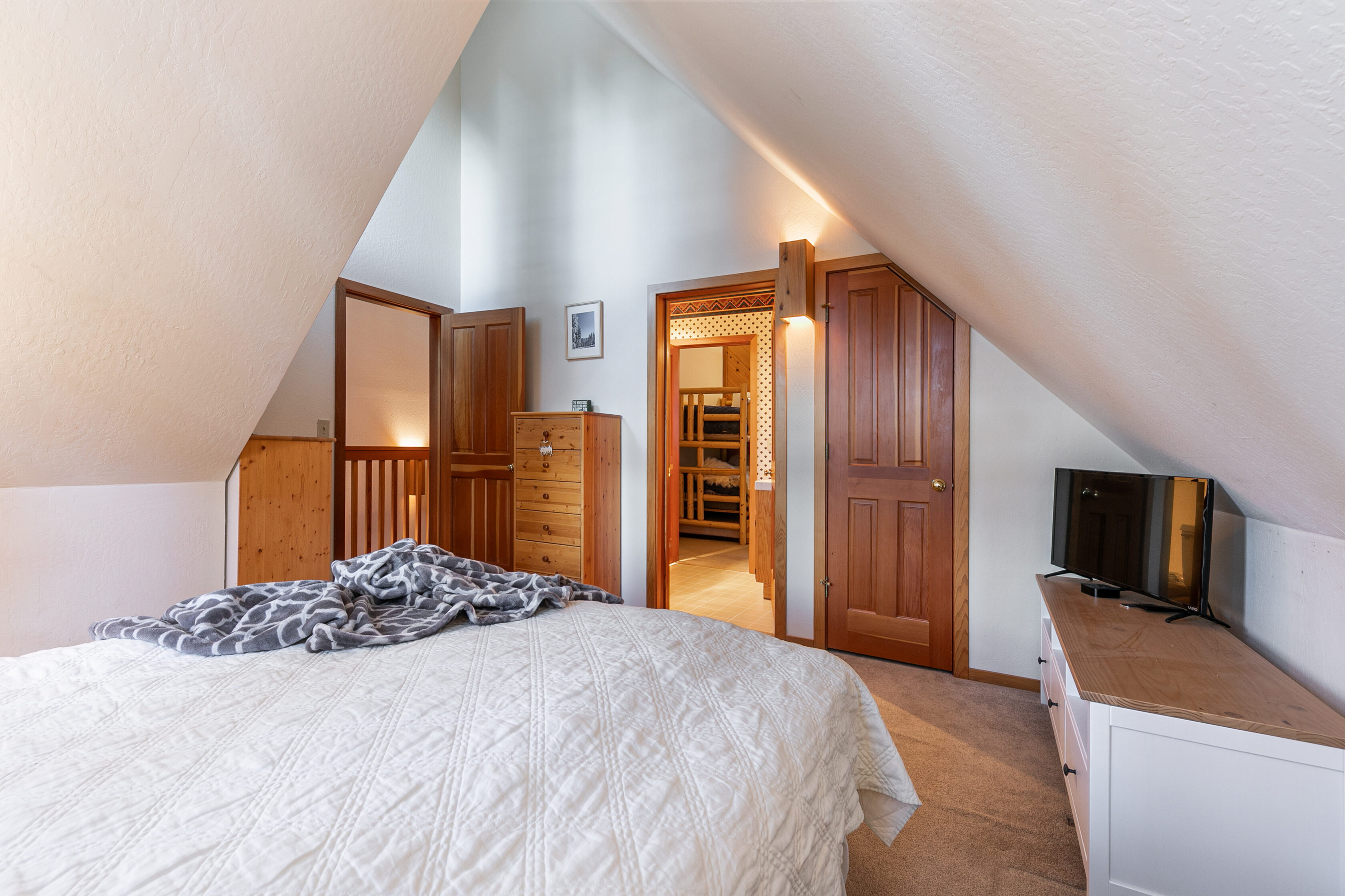 This cozy bedroom features a sloped ceiling and a neutral color palette that emphasizes a warm, rustic aesthetic. It includes a white quilt-covered bed, a wooden dresser, and direct access to an adjoining room with bunk beds, all framed by elegant wooden door trim.