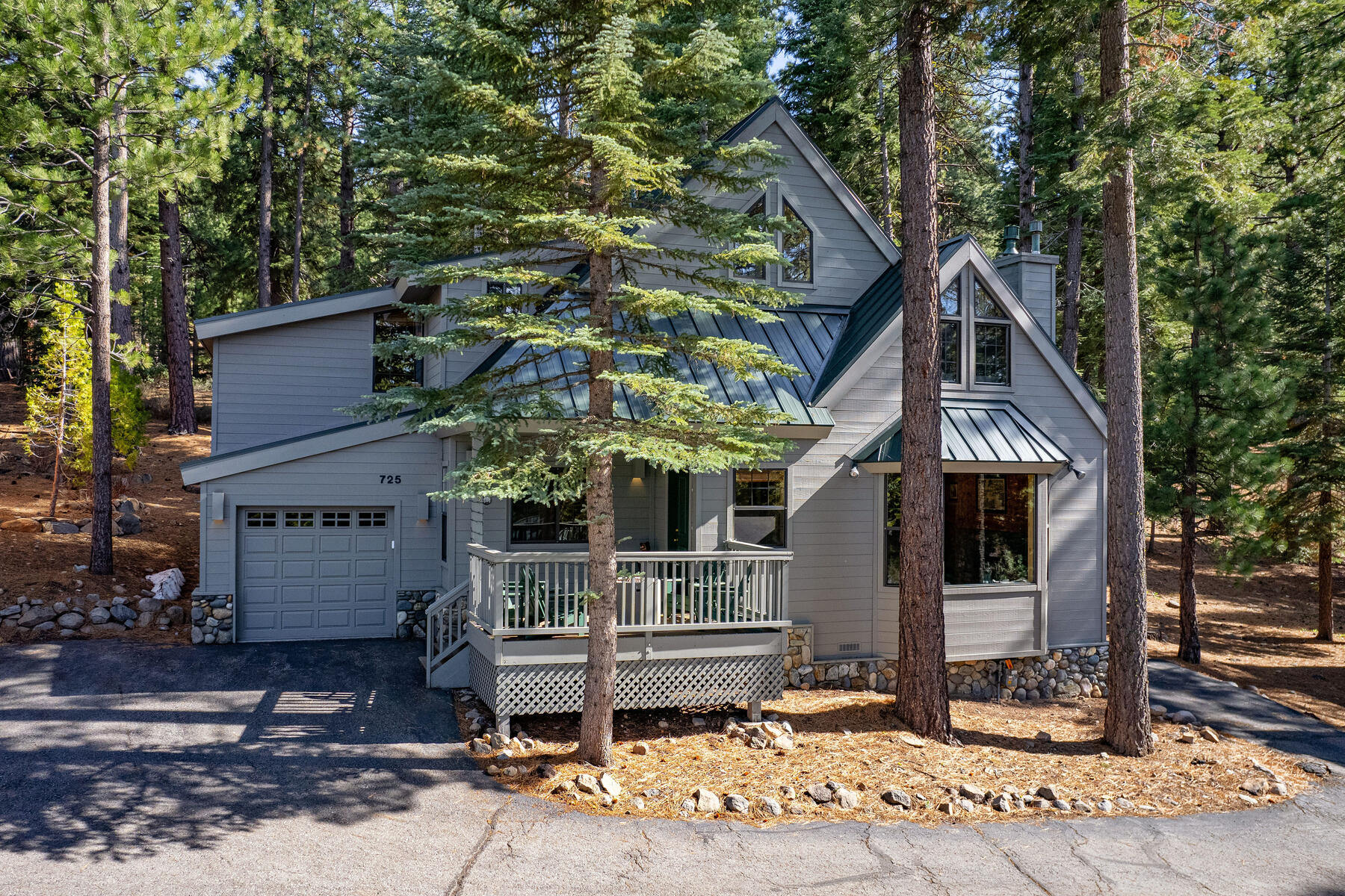 This charming mountain chalet-style home features cool grey siding, a green metal roof, and stone foundation accents that blend seamlessly with the surrounding evergreen trees. The property includes an attached garage, a welcoming wooden front deck, and large windows that invite abundant natural light into the living space. Its rustic yet refined aesthetic makes for an inviting mountainous retreat.