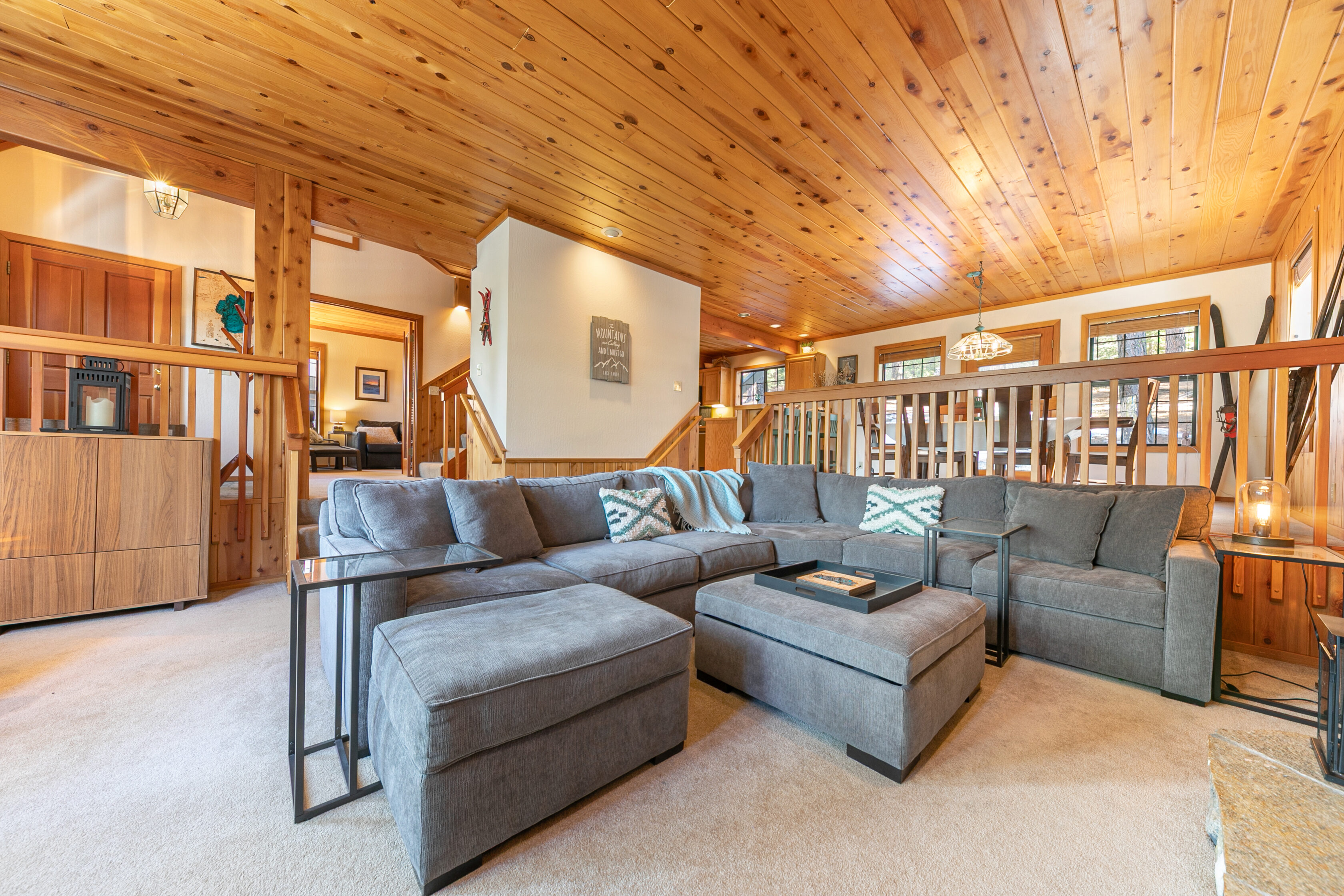 This cozy living room features a spacious sectional sofa with neutral grey upholstery and matching ottomans, ideal for comfortable lounging. The room is characterized by warm wood-paneled ceilings, a rustic open-concept layout, and a split-level design that transitions into a dining area. Decorative touches like patterned throw pillows and wood-accented walls evoke a inviting cabin-style atmosphere.